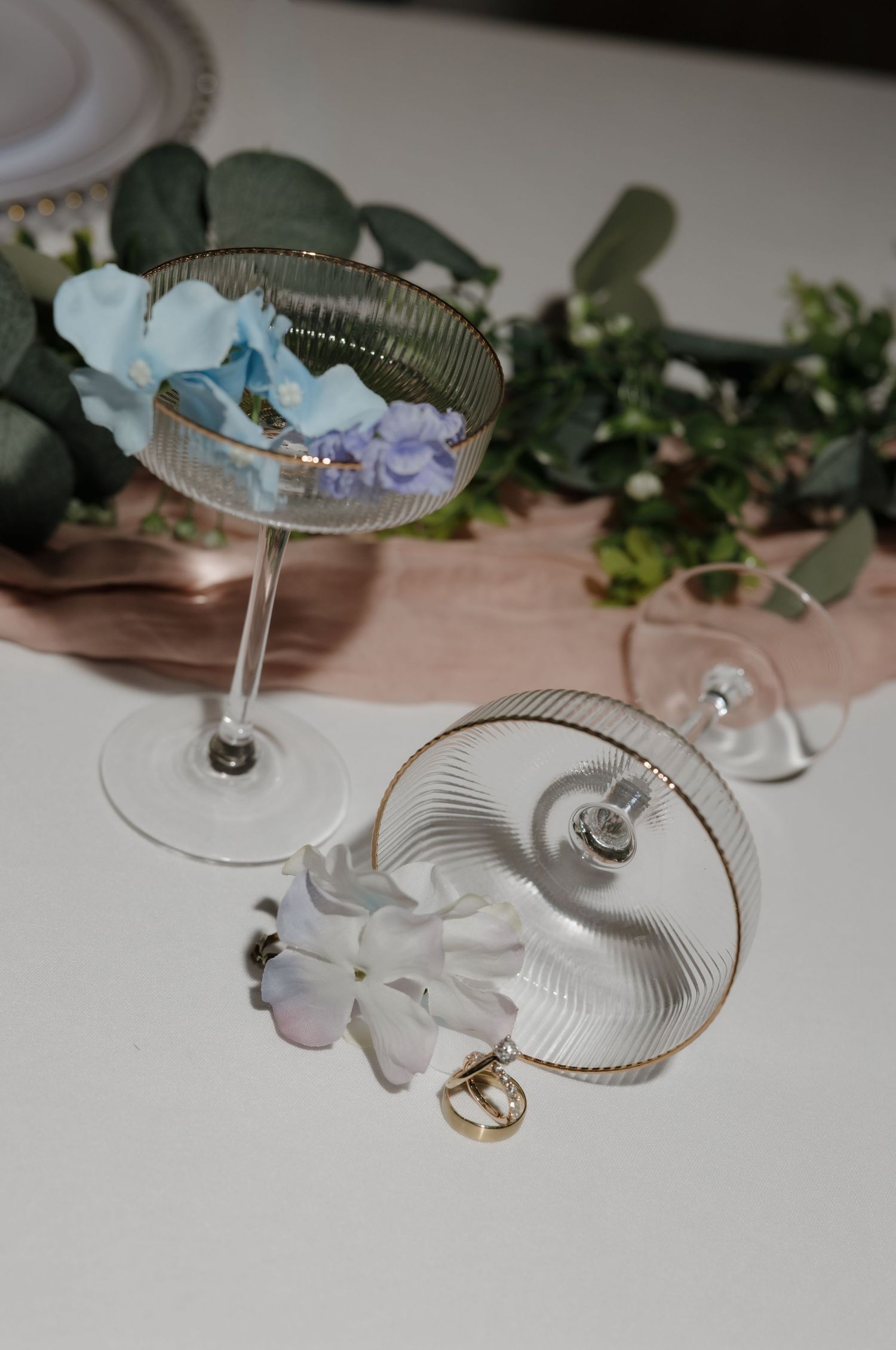 Elegant wedding rings displayed in ribbed crystal glasses with floral decor.