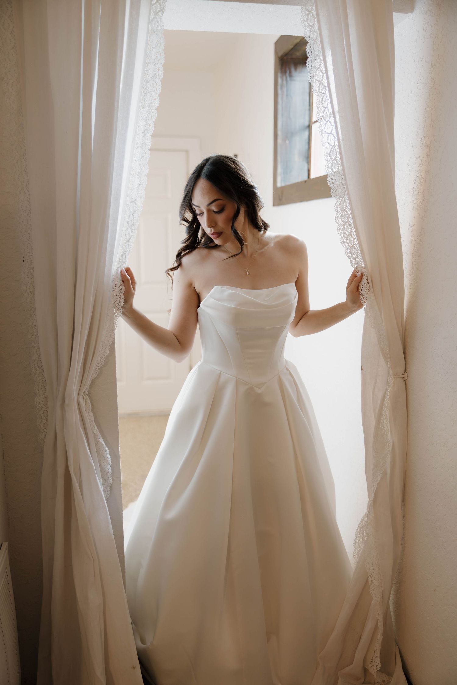 Elegant bride in strapless white ball gown stands between sheer curtains by a sunlit window.