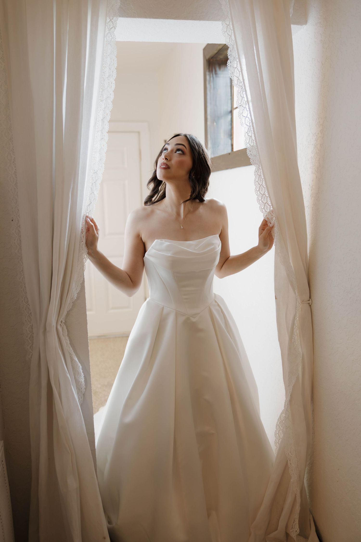 Elegant bride in strapless white ballgown standing between sheer curtains in soft natural light.