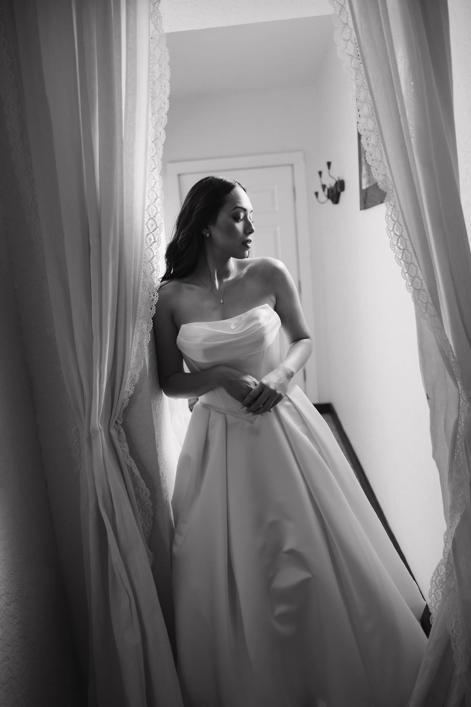 Black and white photo of bride in strapless ballgown gazing out window between lace curtains.