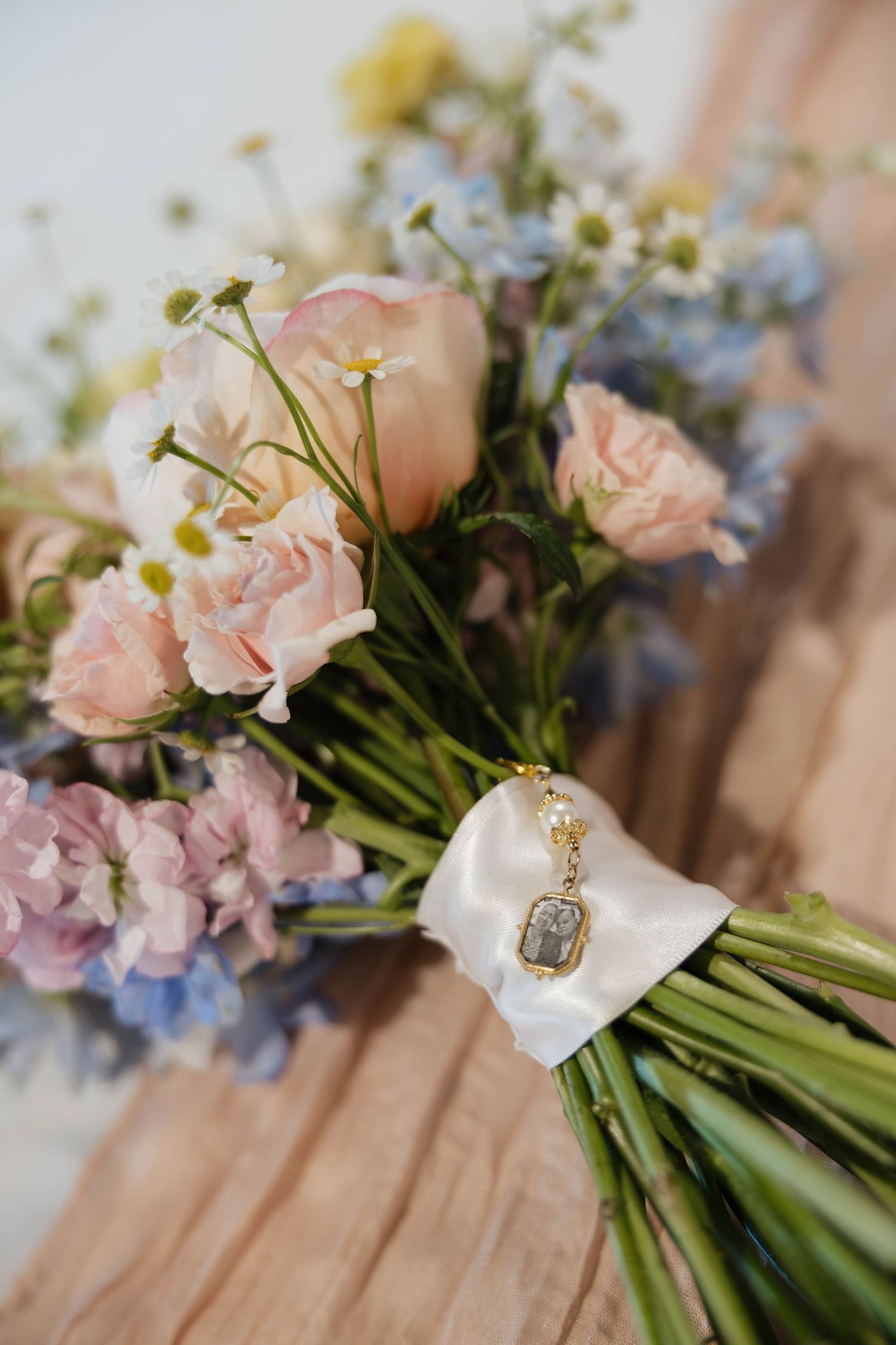 Close-up of a pastel bridal bouquet with pink roses, blue flowers, and a gold locket charm tied with white ribbon.