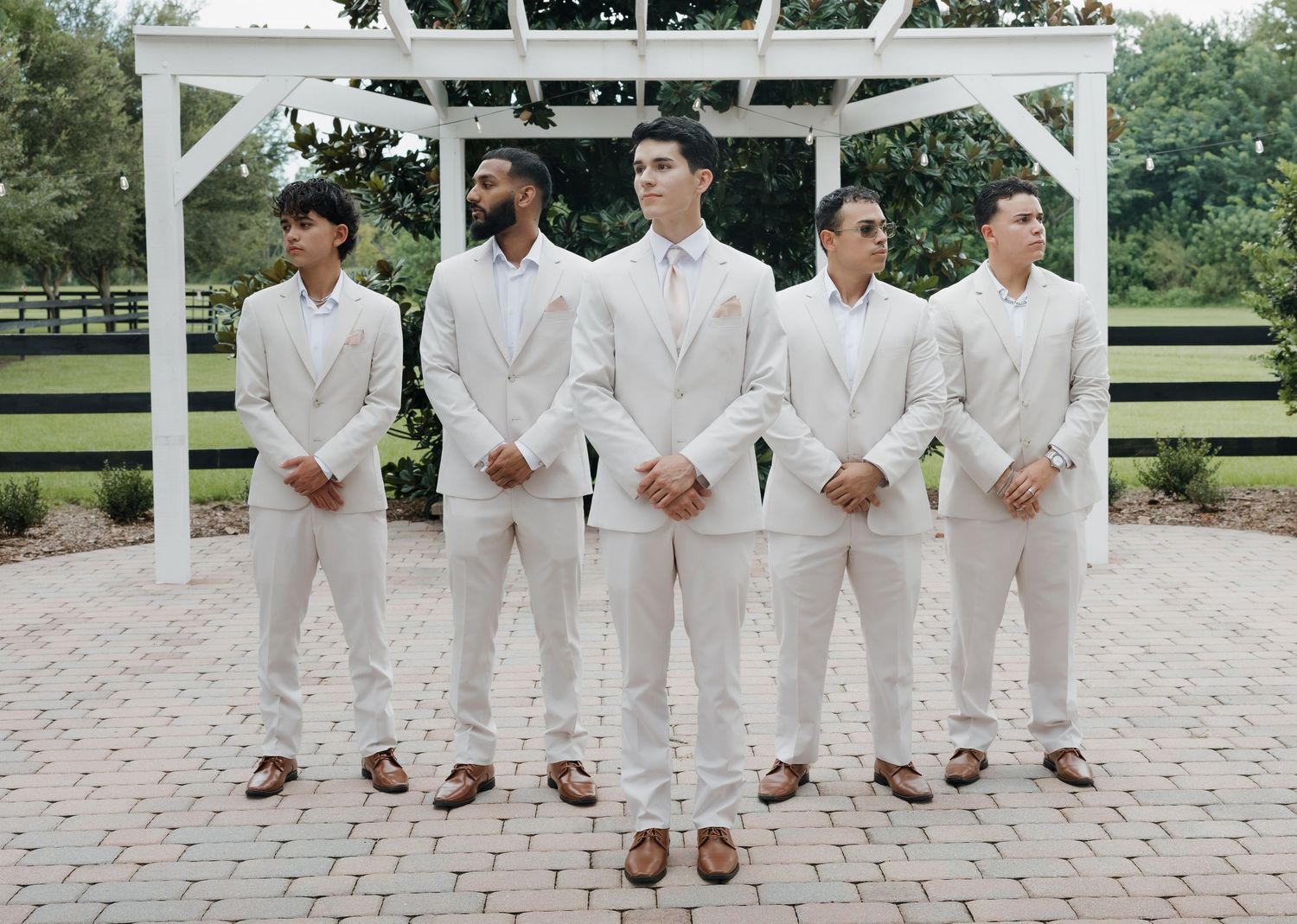 Five groomsmen in matching white suits and brown dress shoes pose outdoors near a white pergola at a wedding.