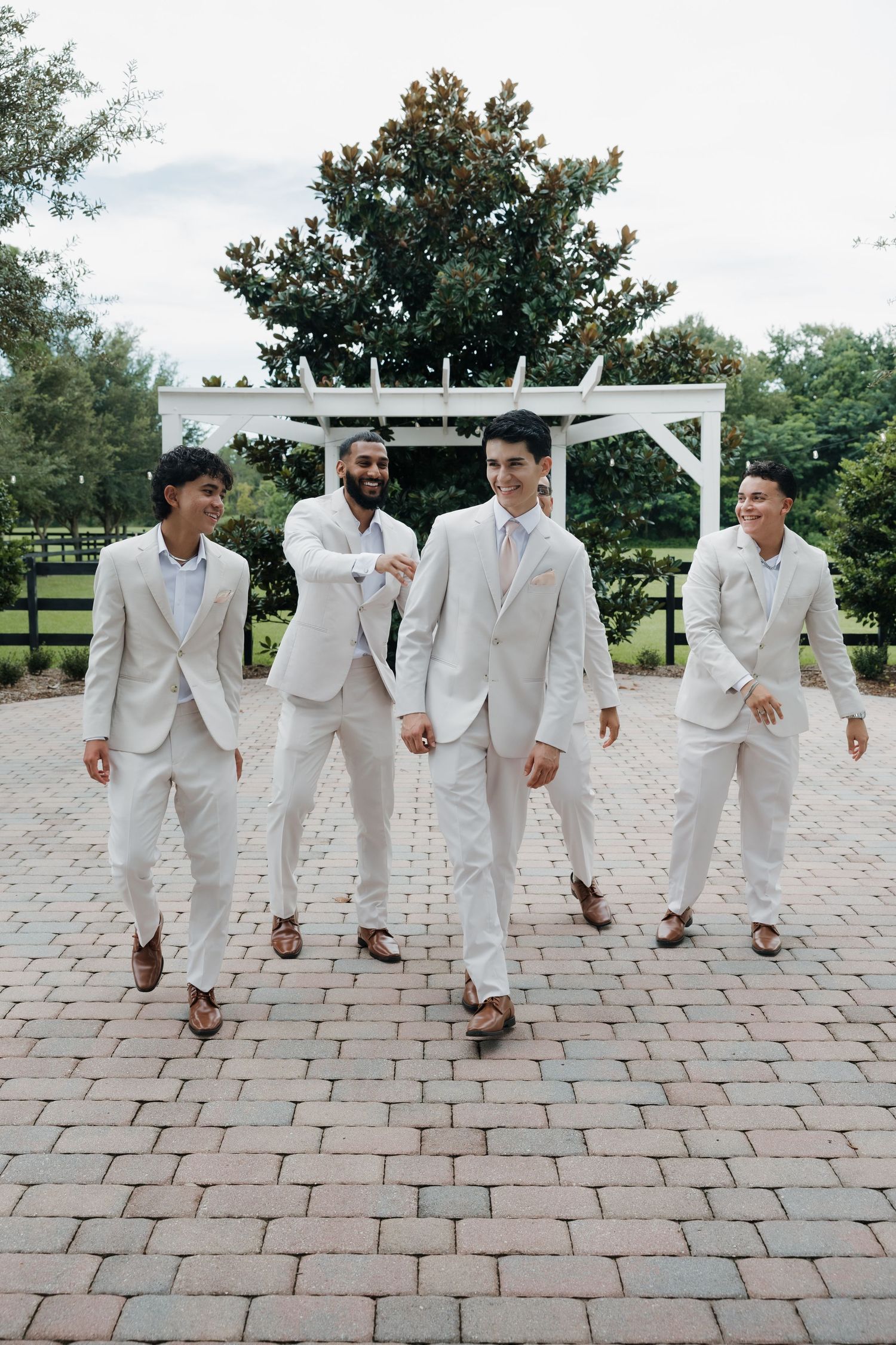 Groom and groomsmen in matching cream suits walking confidently on a brick patio at an outdoor venue.