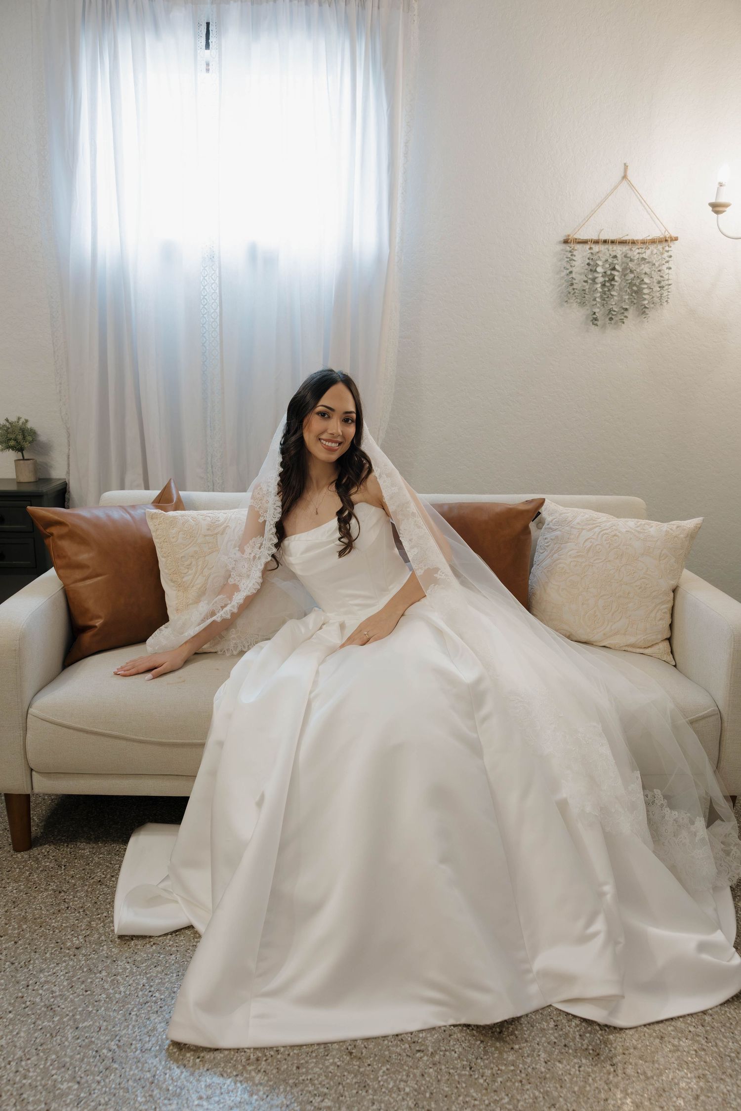 Radiant bride in white gown and veil seated on couch near bright window.