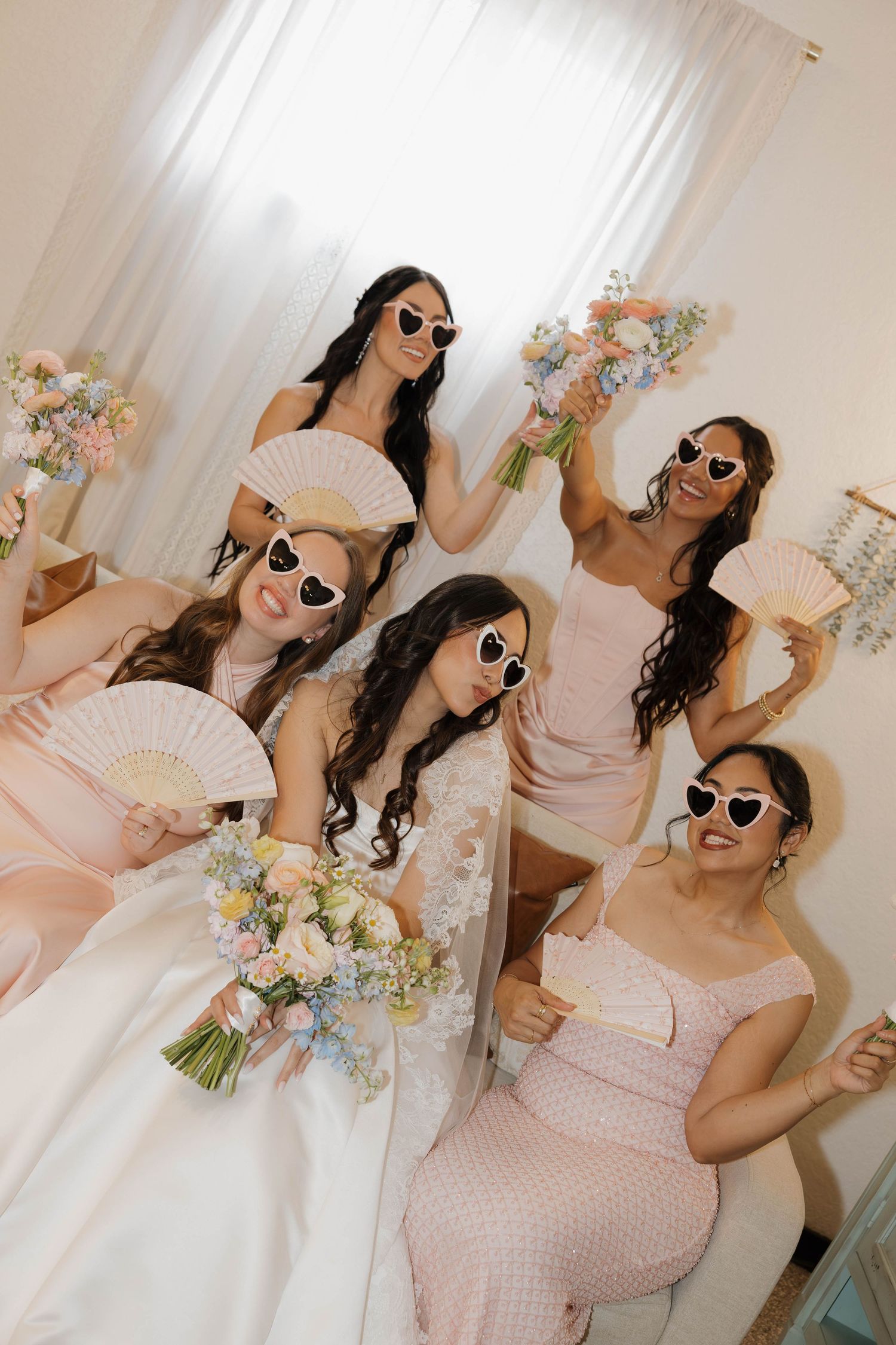 Bride and bridesmaids in blush pink dresses wearing heart-shaped sunglasses holding bouquets and fans in a bright room.
