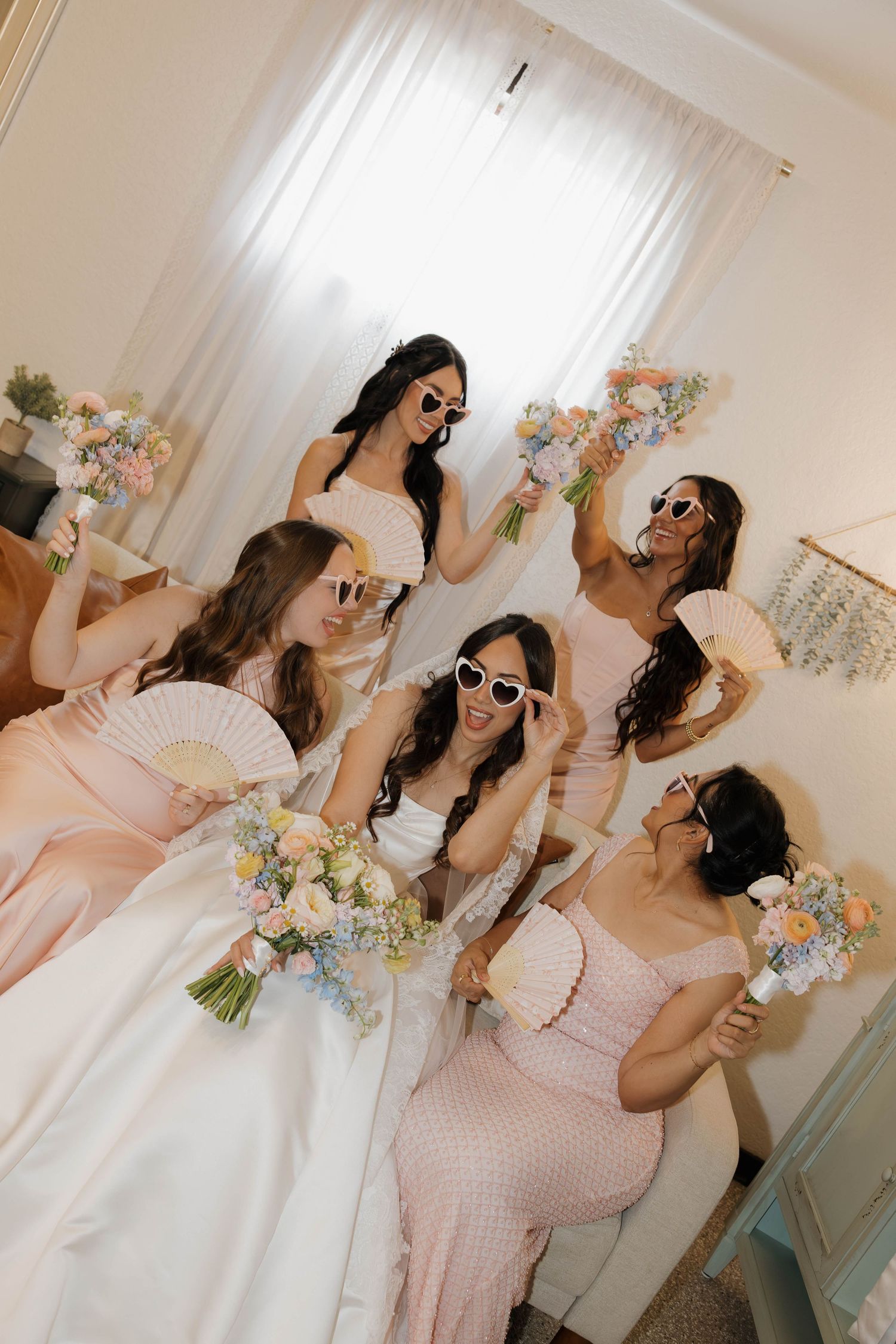 Bride in white gown with bridesmaids in pink dresses posing with heart sunglasses and colorful floral bouquets indoors.