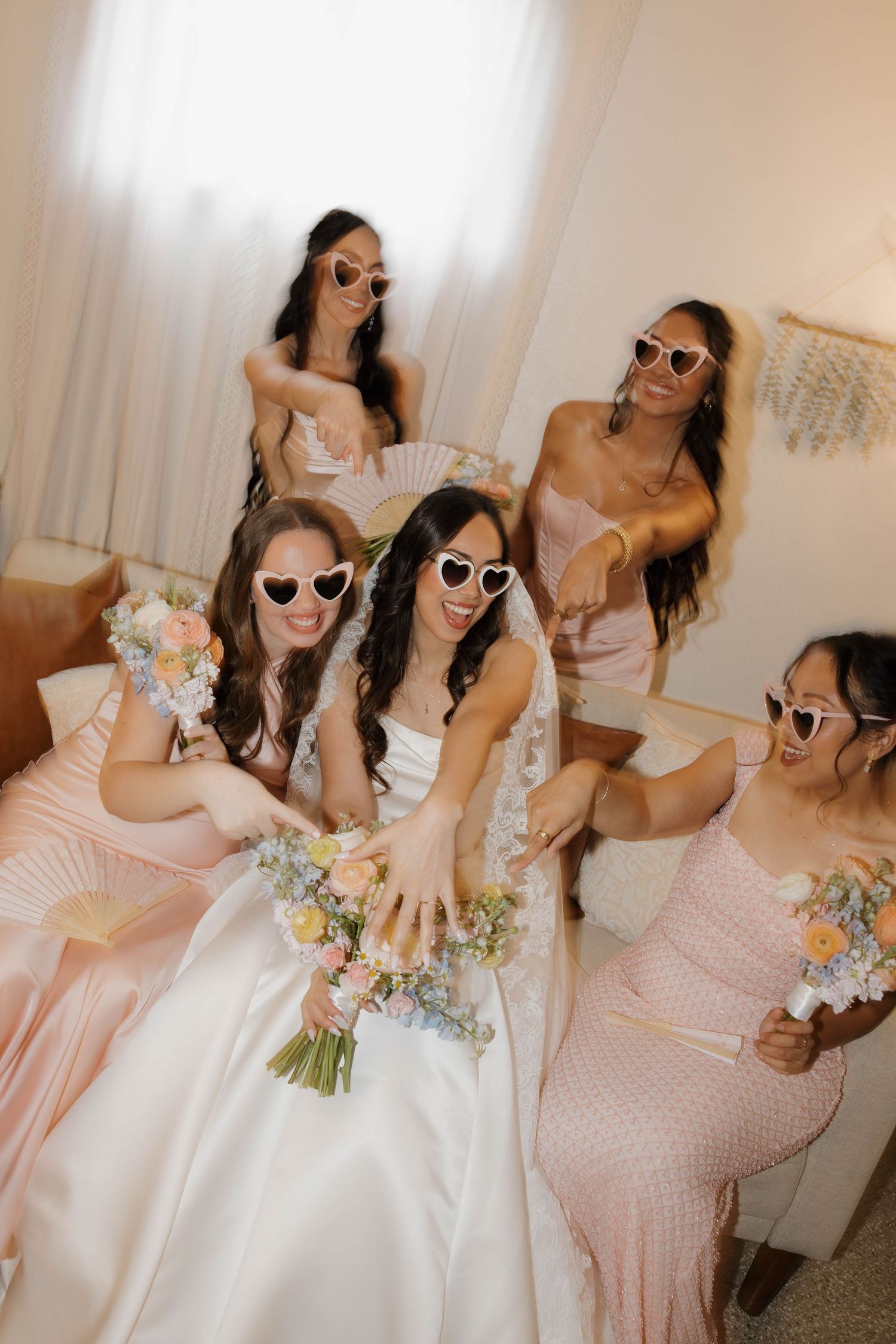 Bride and bridesmaids in pink dresses wear heart sunglasses while showing off wedding ring indoors.