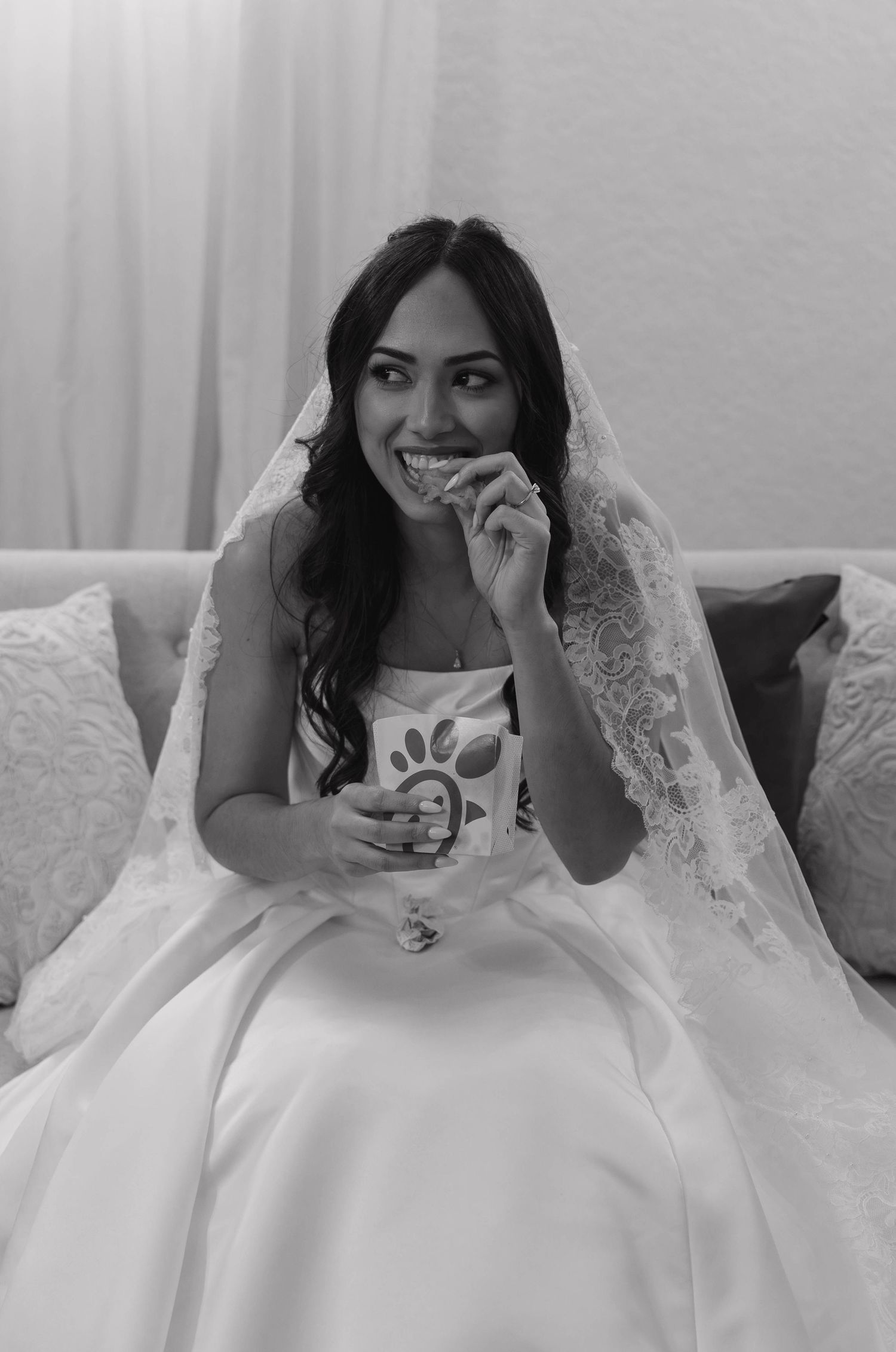 Black and white photo of bride in wedding gown and veil eating Chick-fil-A on a sofa.