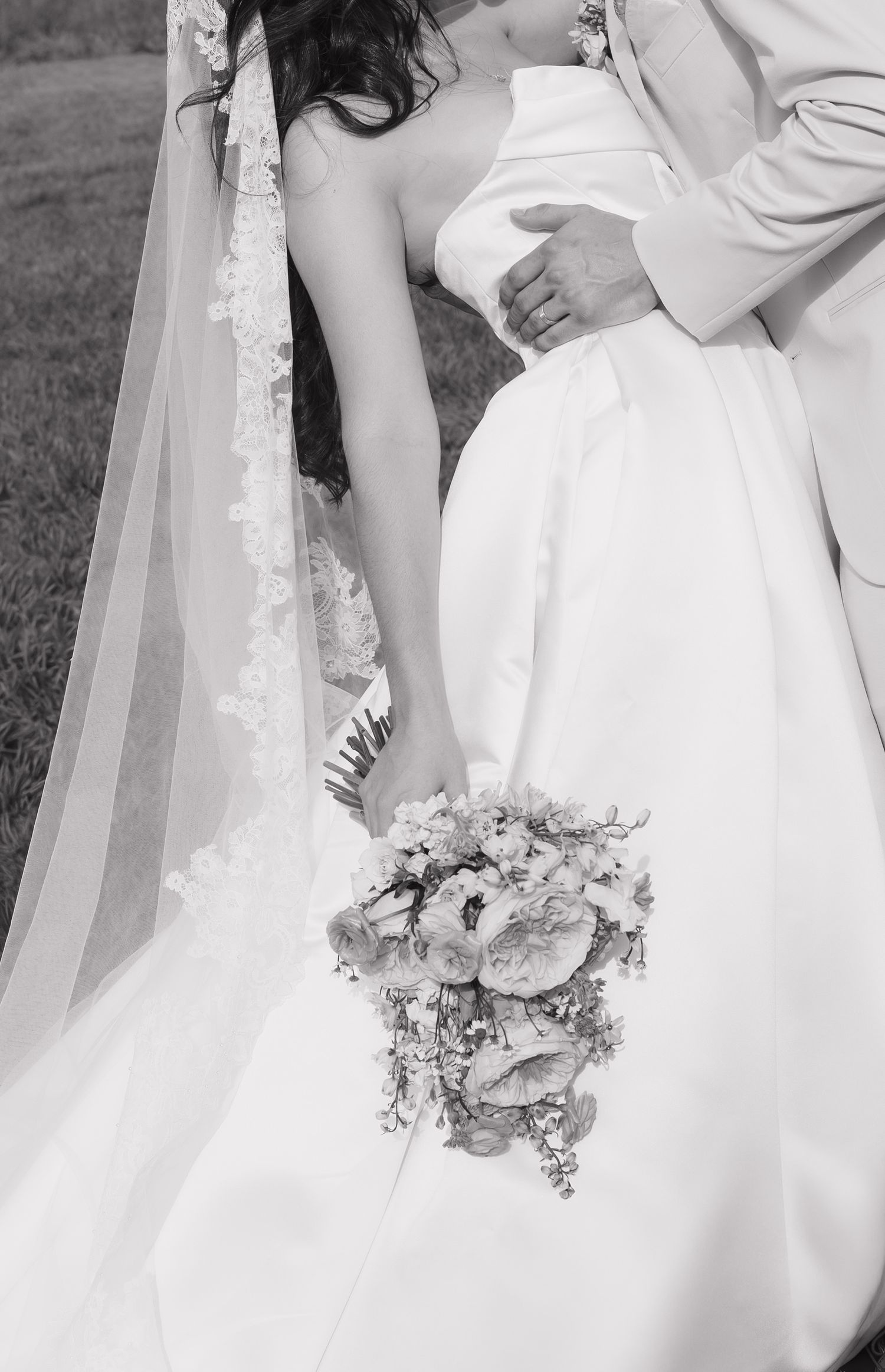 Black and white photo of bride in strapless gown with lace veil holding a cascading floral bouquet.