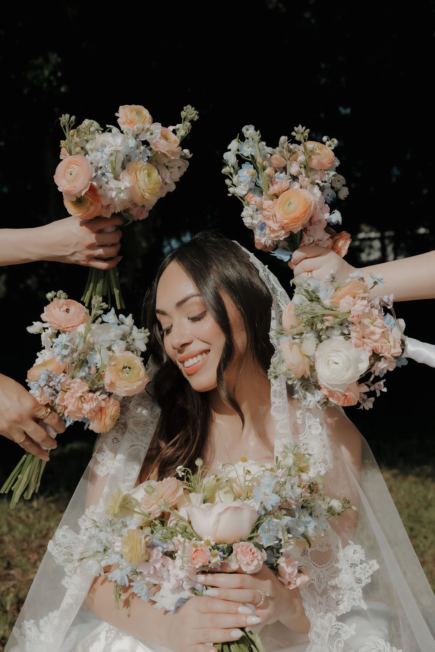 Smiling bride surrounded by pastel pink and blue floral bouquets held by bridesmaids outdoors.
