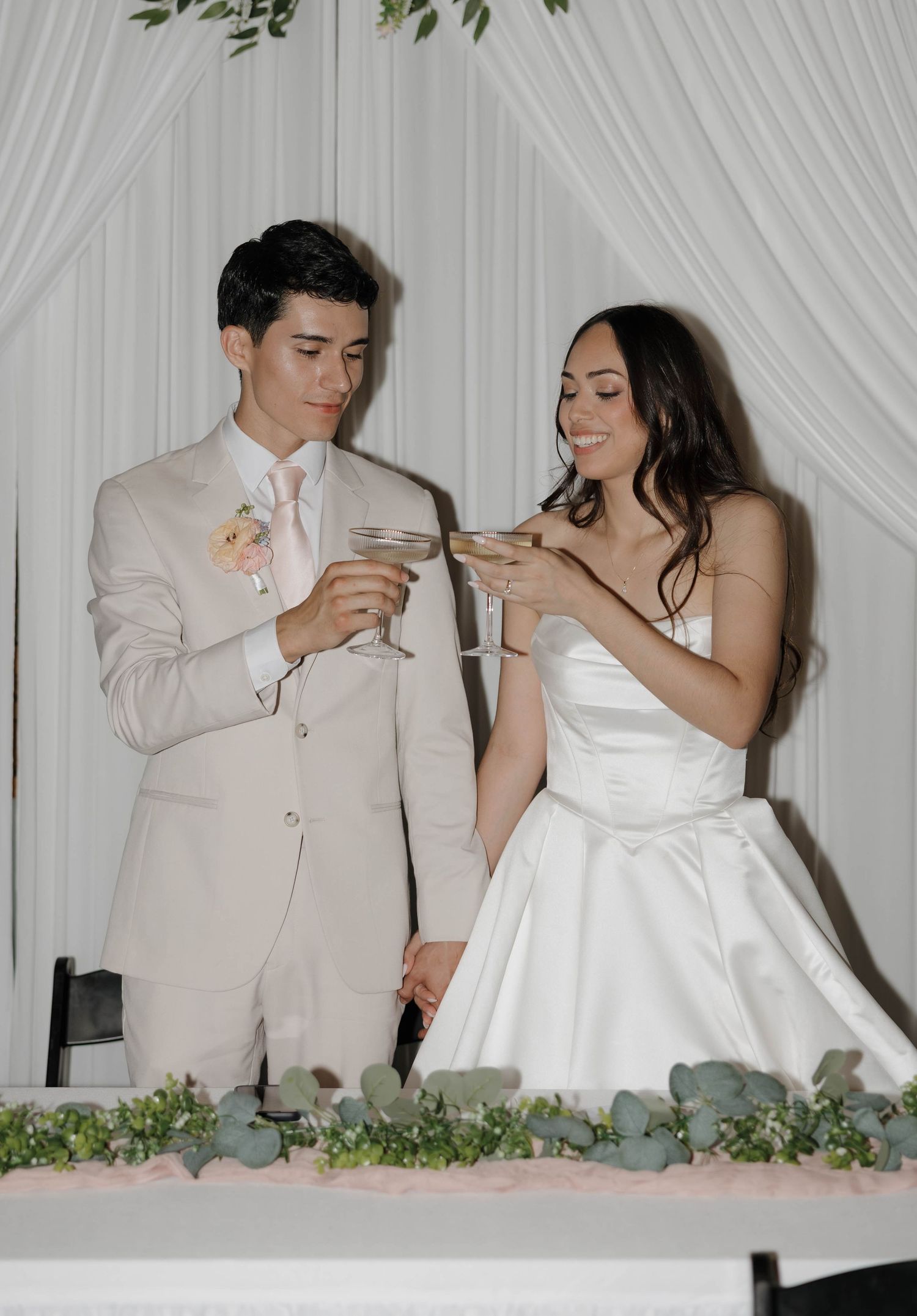 Bride and groom in white wedding attire toasting with champagne glasses at their elegant reception.