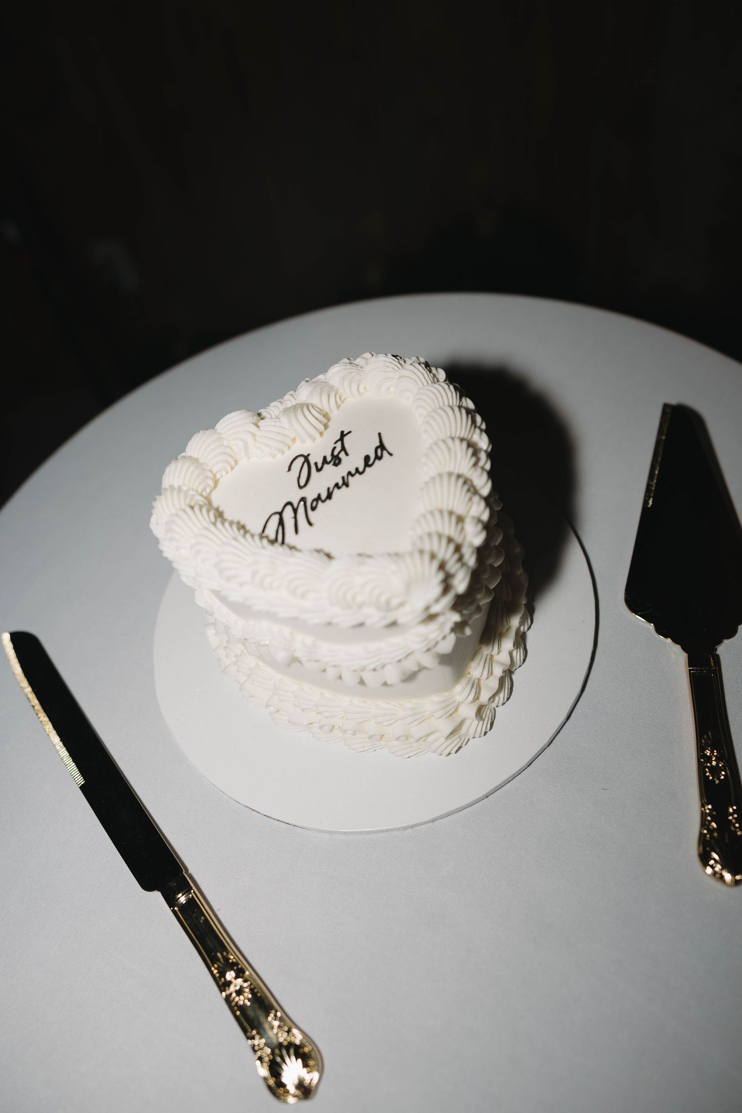 Heart-shaped white wedding cake inscribed with Just Married, displayed on a round board with gold cake cutting utensils.