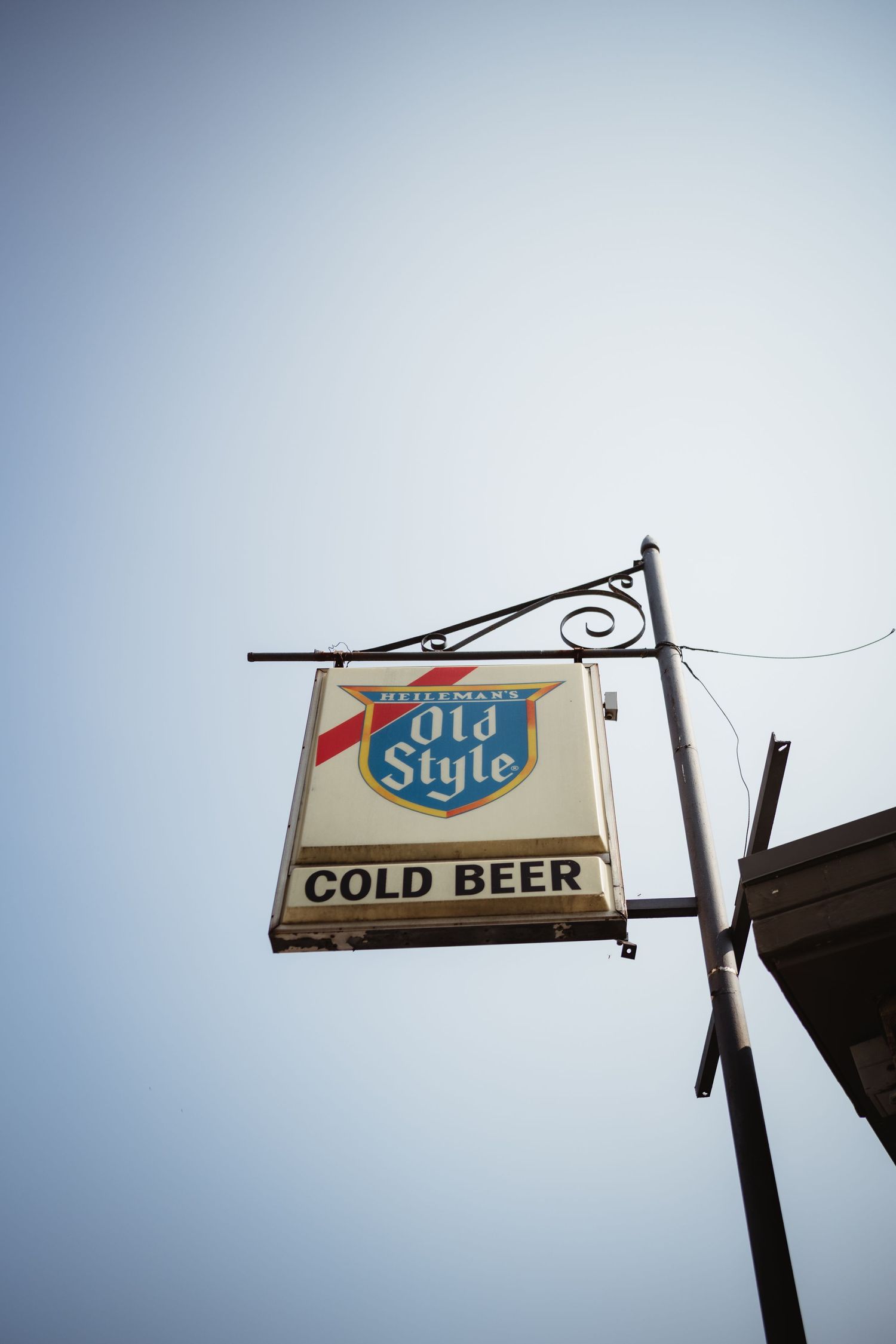 Vintage Old Style Cold Beer advertising sign mounted on a decorative metal bracket against a clear blue sky.