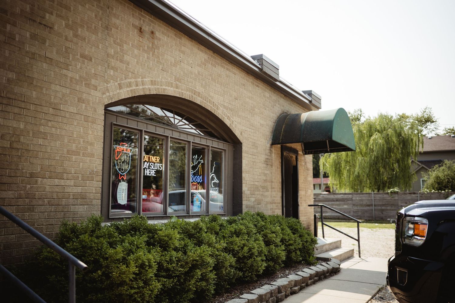 Brick storefront with arched window display, green awning, and lush weeping willow tree in background on sunny day.