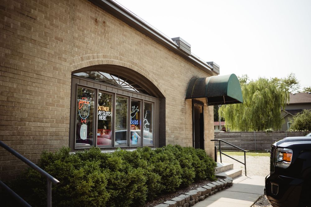 Brick storefront with arched window display, green awning, and lush weeping willow tree in background on sunny day.