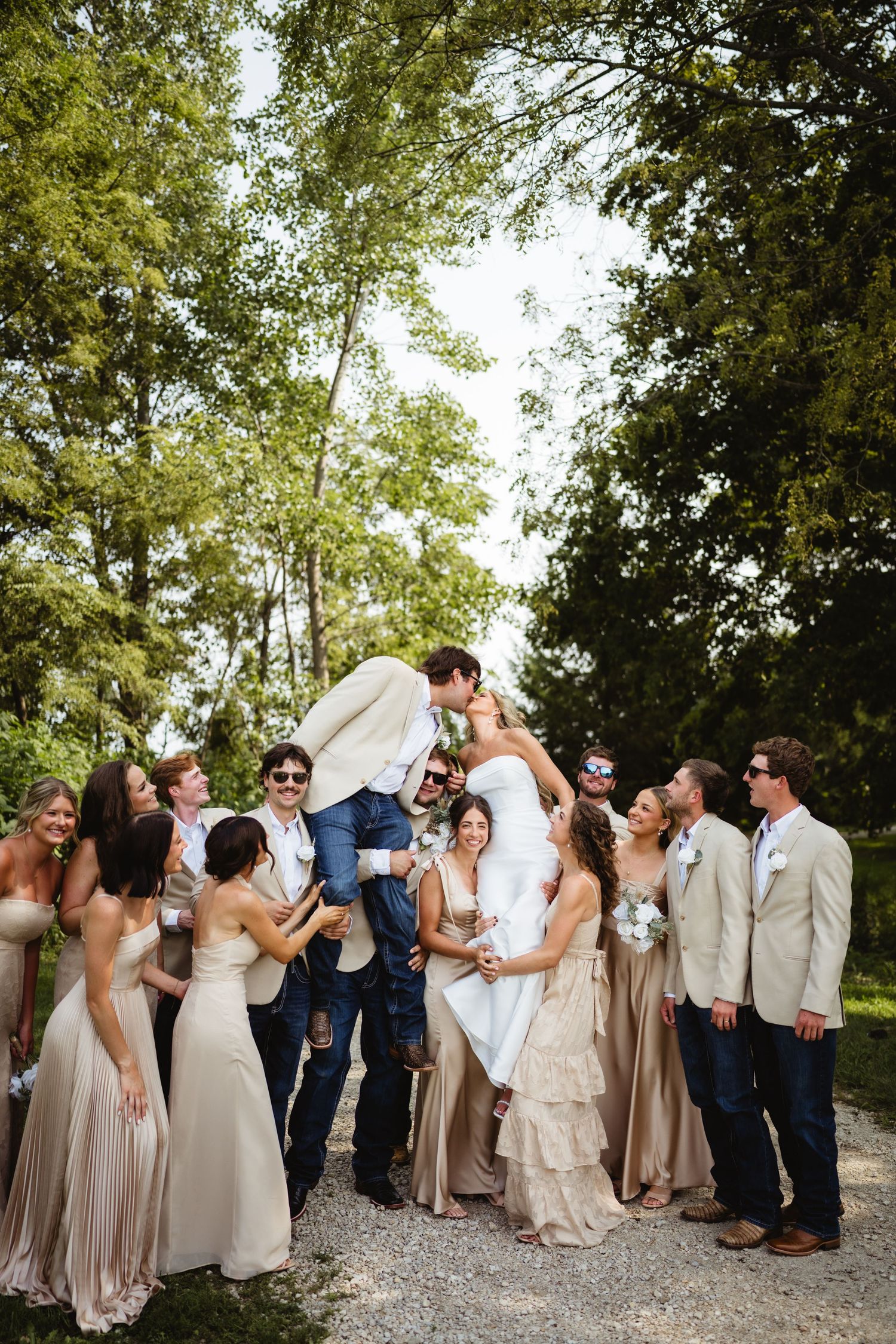 Wedding party celebrates outdoors as groom lifts bride, surrounded by bridesmaids in champagne gowns and groomsmen in tan suits.