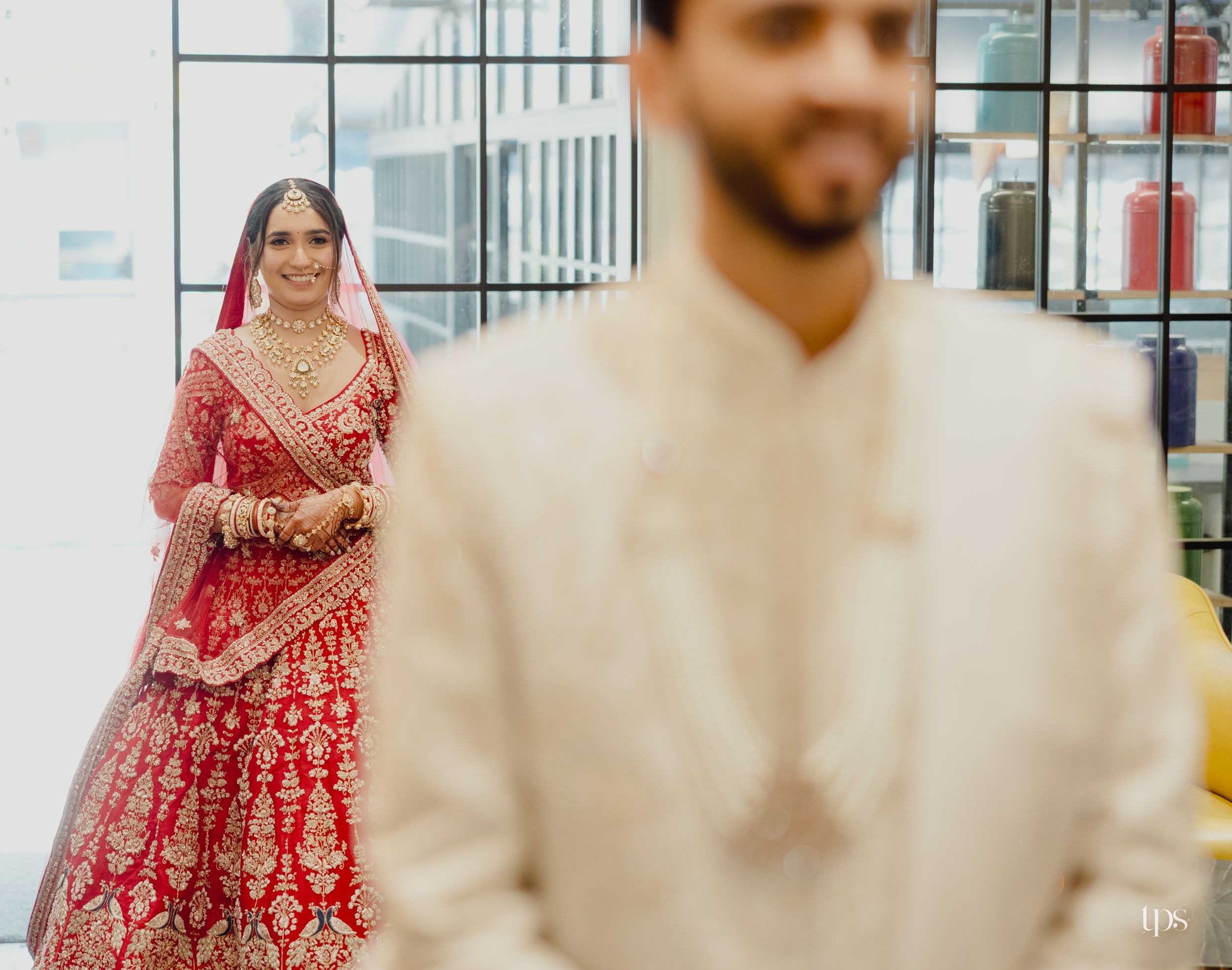 Indian bride in red bridal lehenga stands behind groom in cream sherwani during traditional wedding ceremony.