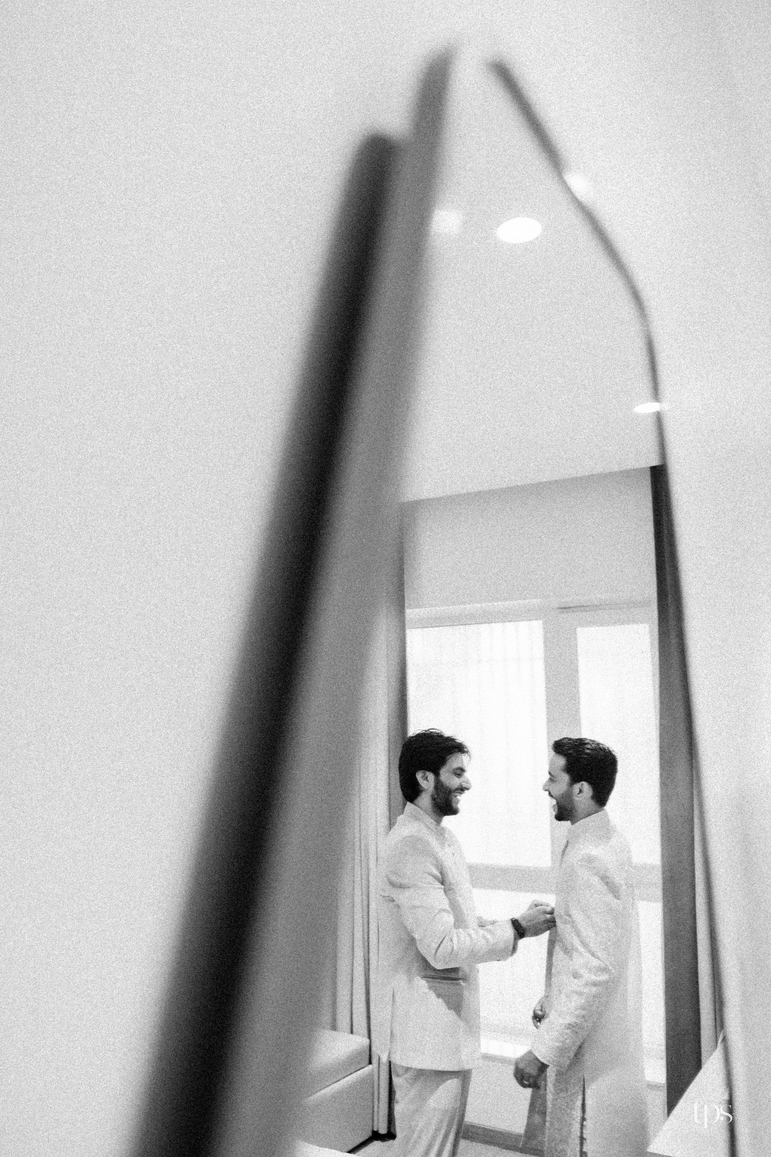 Black and white photo of two grooms in white suits sharing a moment, reflected in a tall mirror.