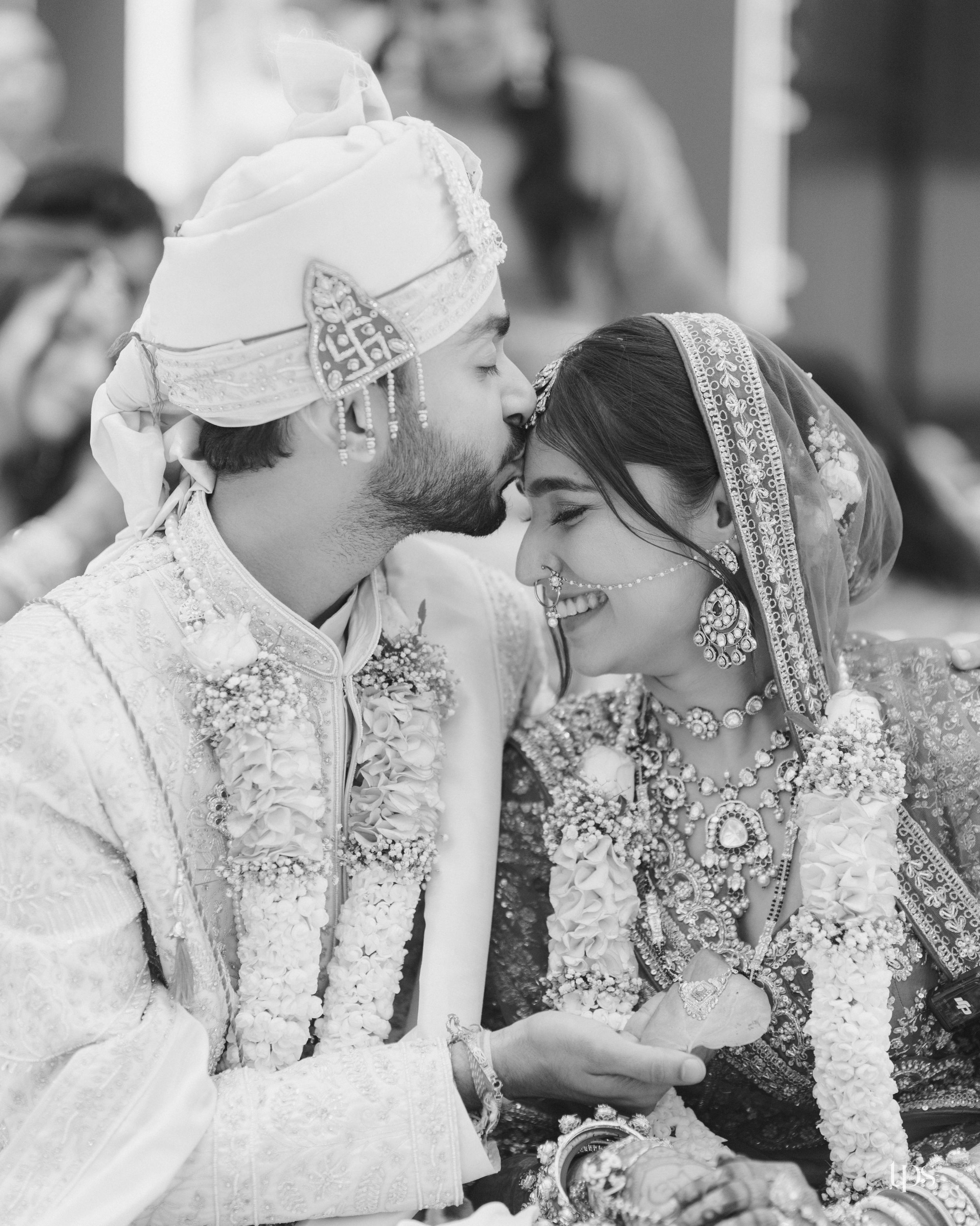 Black and white photo of a groom kissing bride on cheek during traditional Indian wedding ceremony with floral garlands.