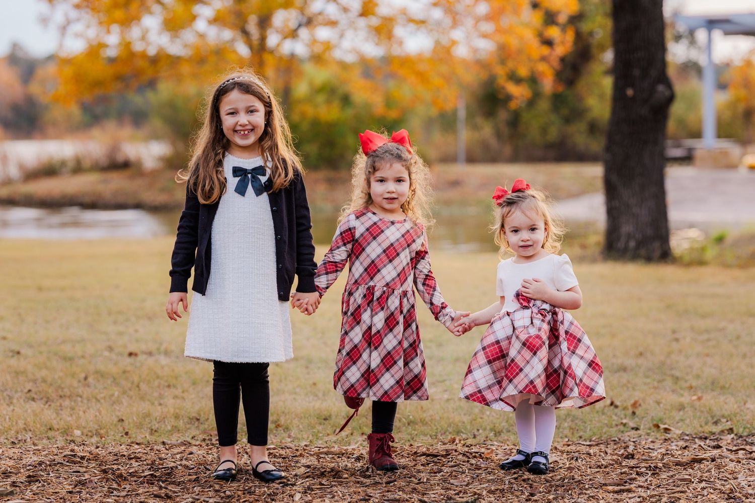 Three young girls in holiday dresses hold hands smiling in a park with autumn foliage backdrop.