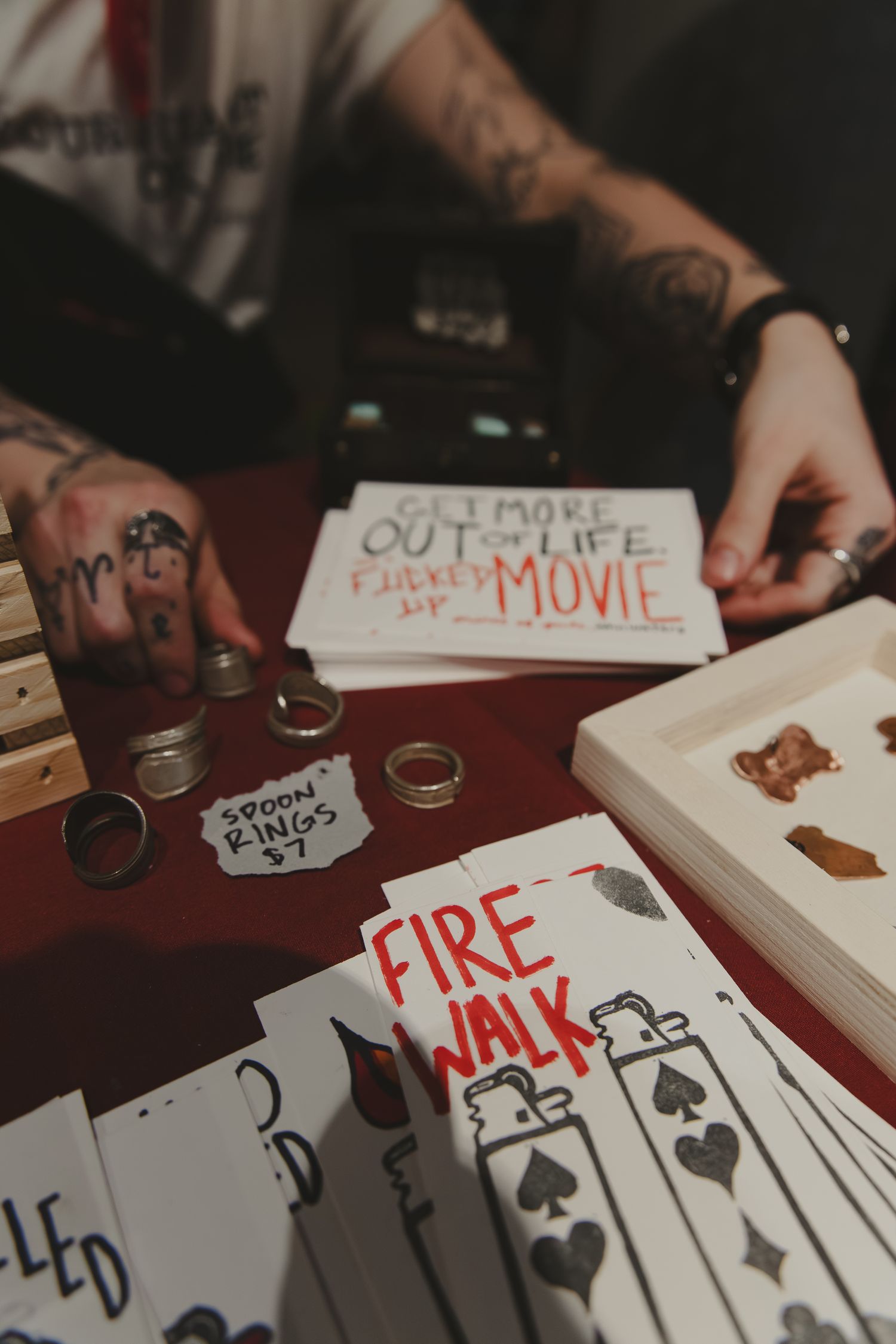 Tattooed hands arranging merchandise including spoon rings and Fire Walk zine cards on a red table at an indie market.
