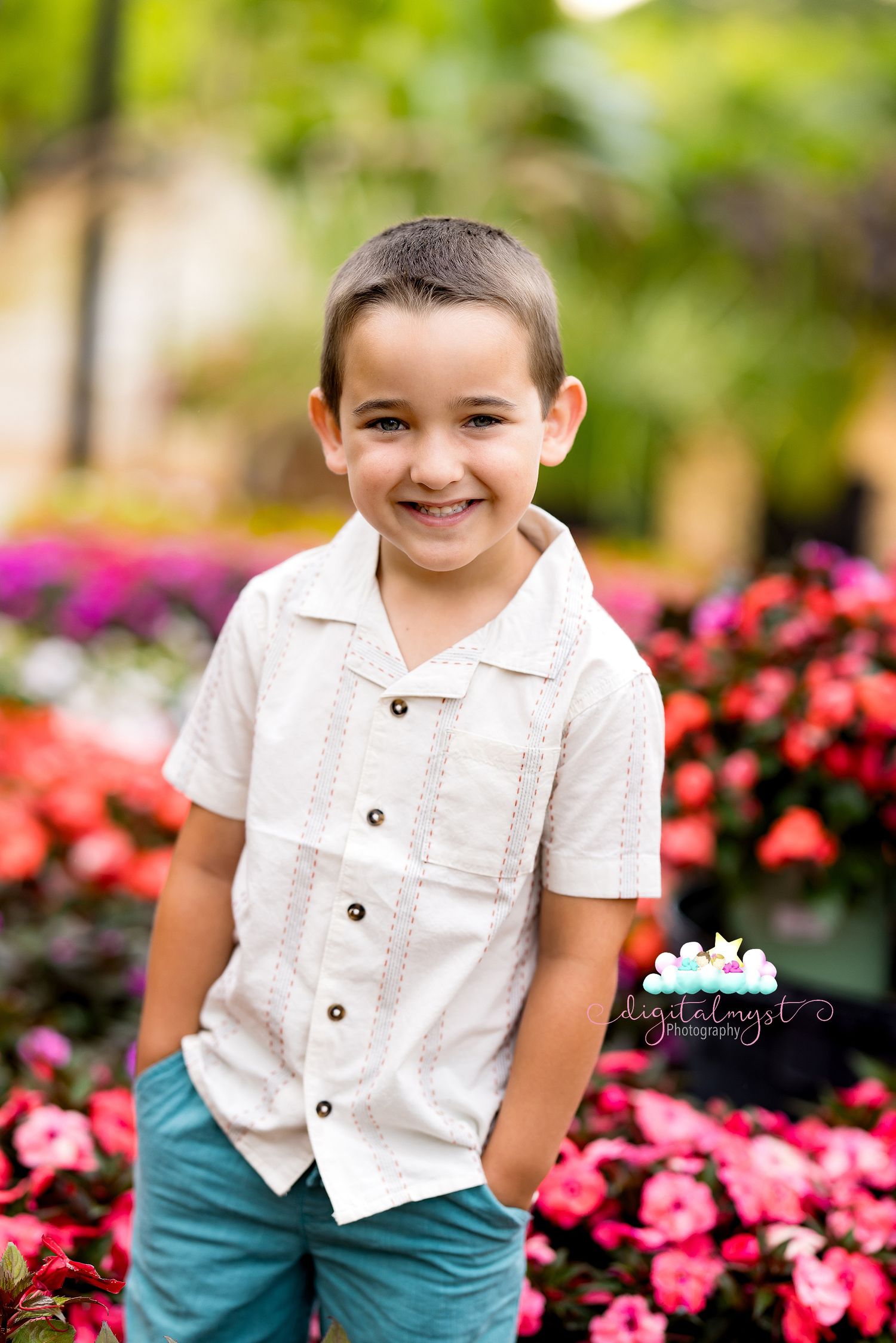 A smiling young boy in a white button-up shirt and teal pants poses in front of colorful flowers.