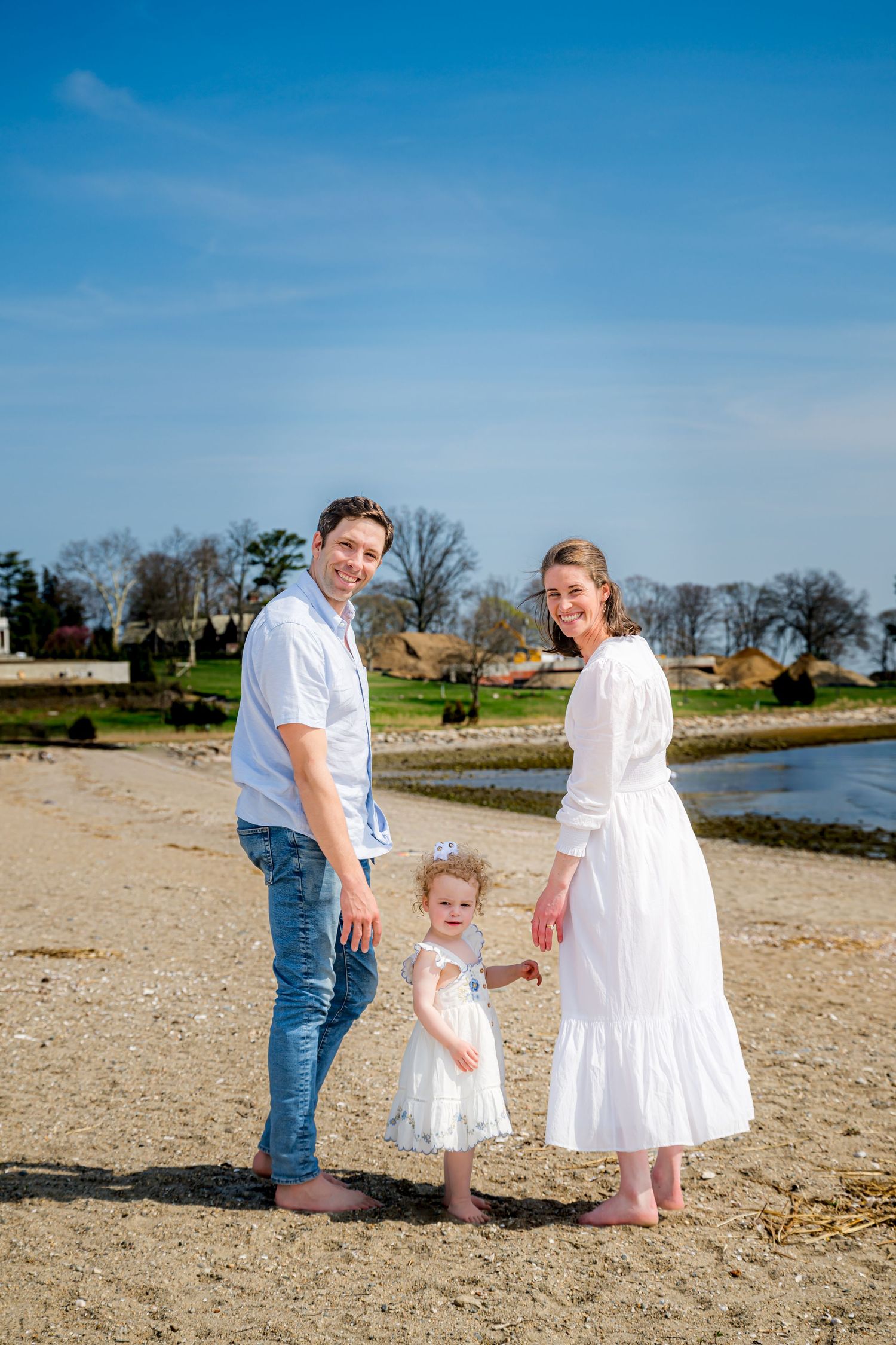 Family of three posing on sandy beach, parents in white outfits with toddler daughter, blue sky backdrop.