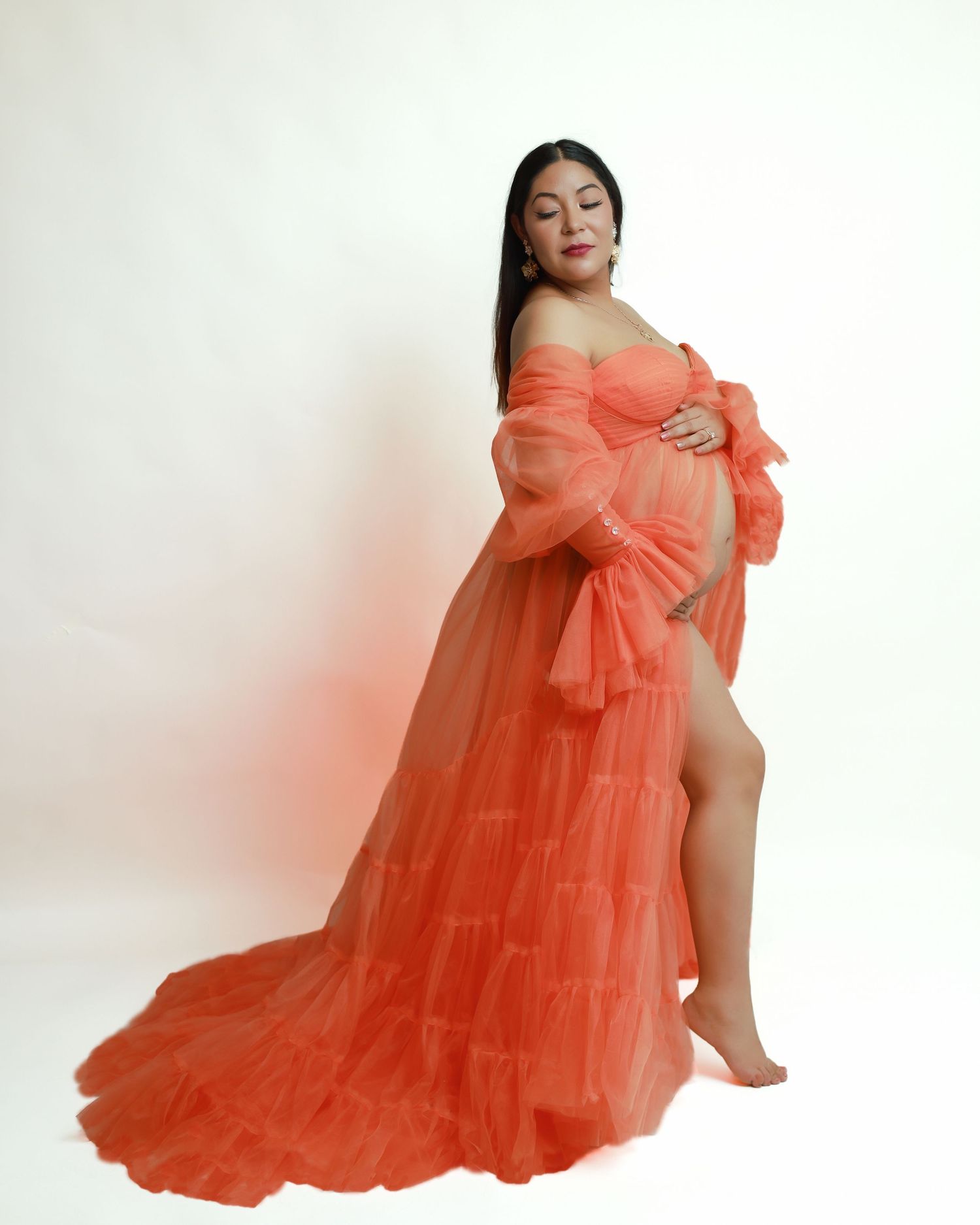 Pregnant woman posing in an elegant off-shoulder coral tulle maternity gown with long sleeves and a flowing train.