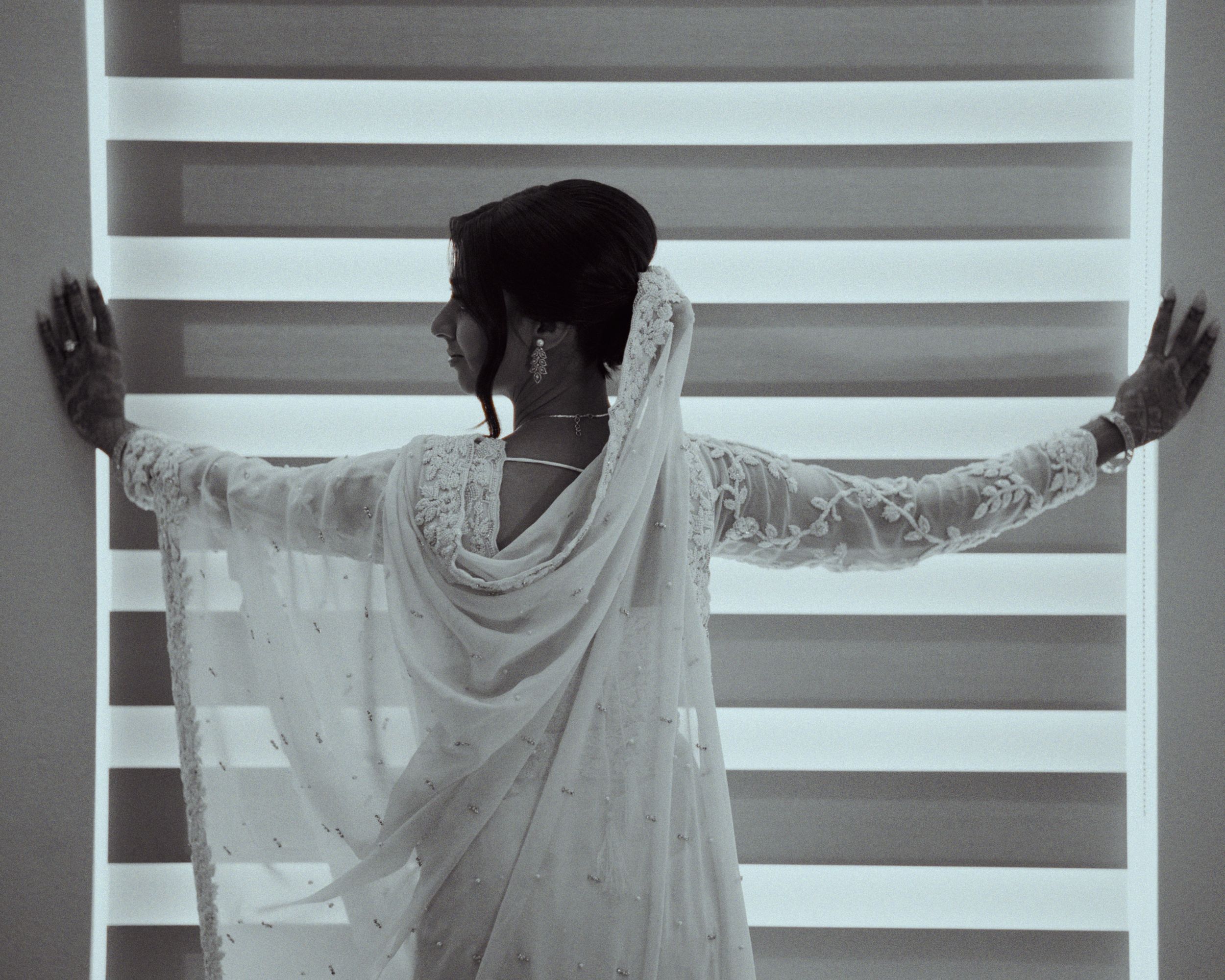 Ismaili bride white embroidered saree with arms outstretched against striped background, black and white photo.