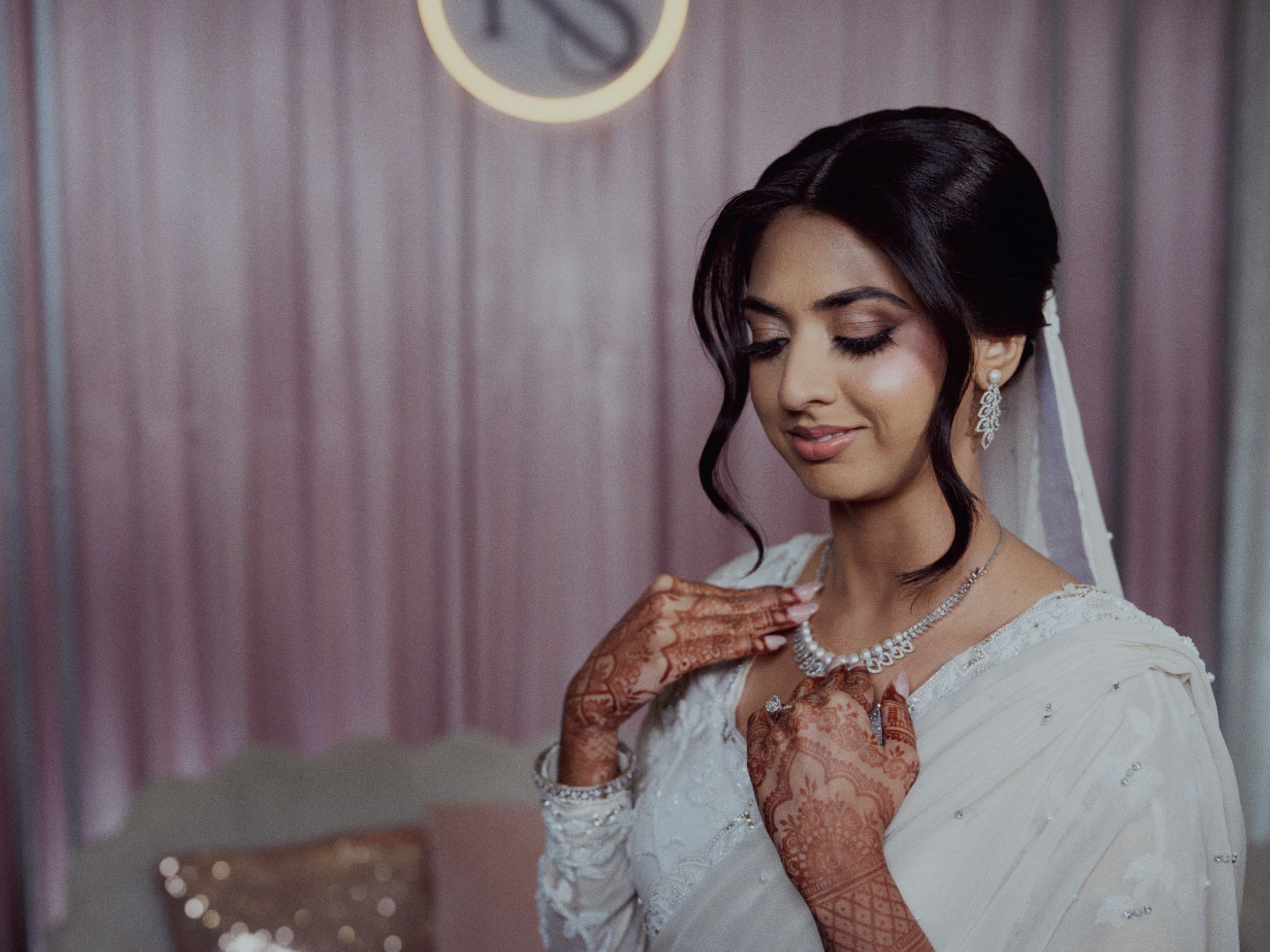 South Asian bride in white outfit with henna hands adjusting diamond necklace against pink backdrop.