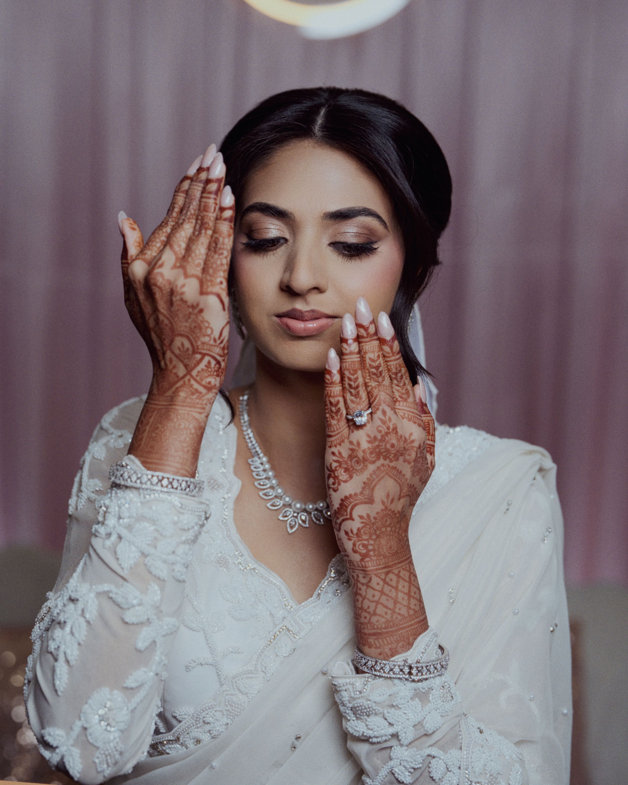 South Asian bride in white outfit with intricate henna designs on hands, wearing diamond jewelry.