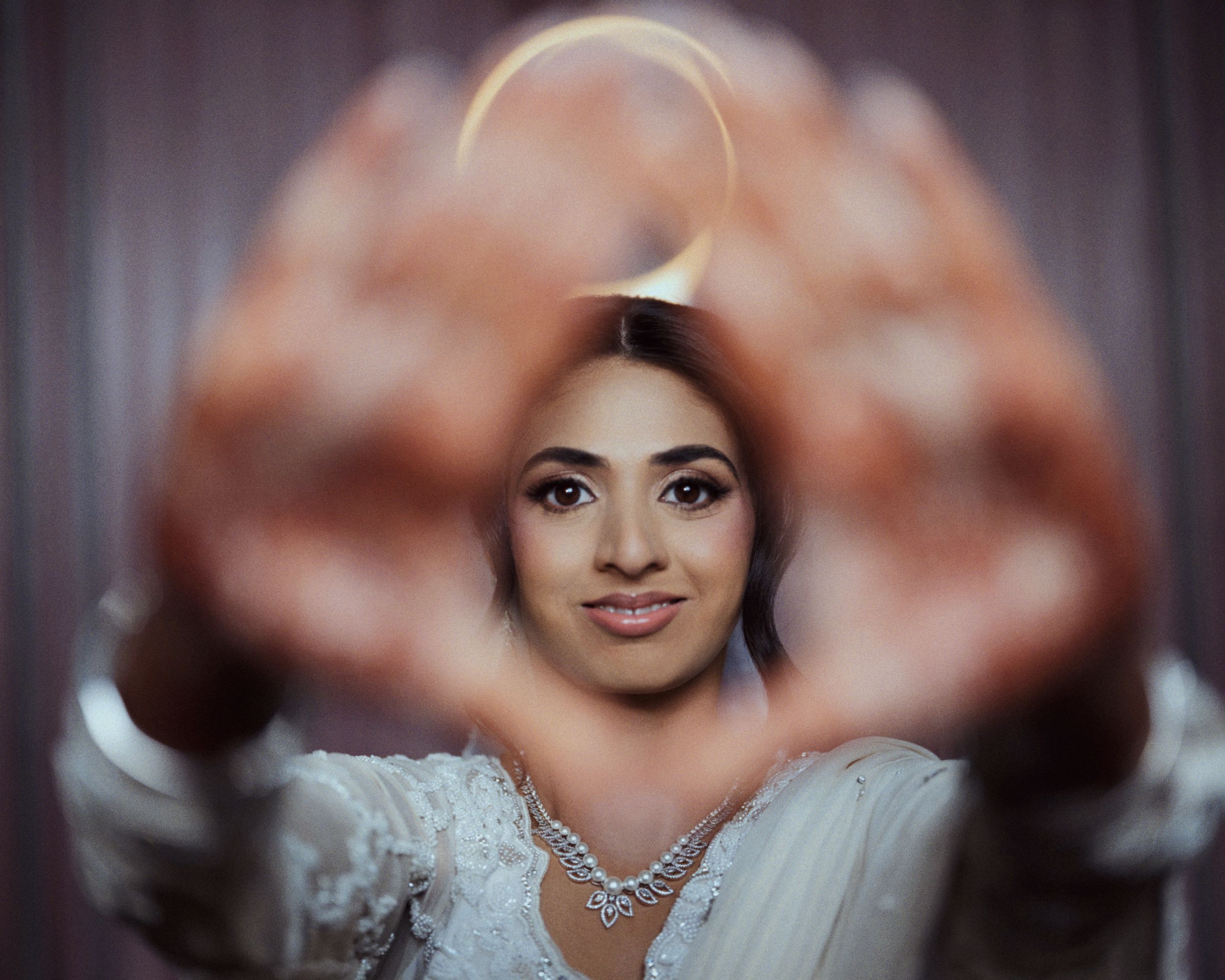 Bride in white lace gown frames glowing ring with hands, creating bokeh effect in elegant wedding portrait.
