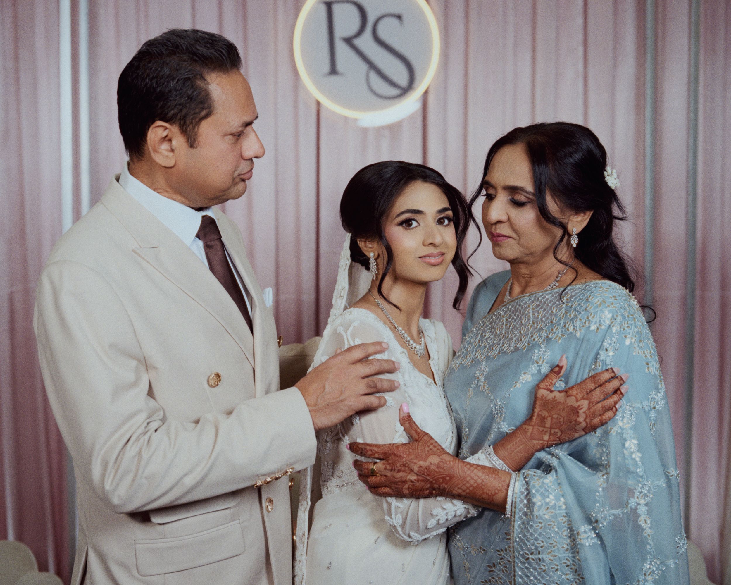 A bride in white with henna stands between her parents in elegant attire at a wedding ceremony.