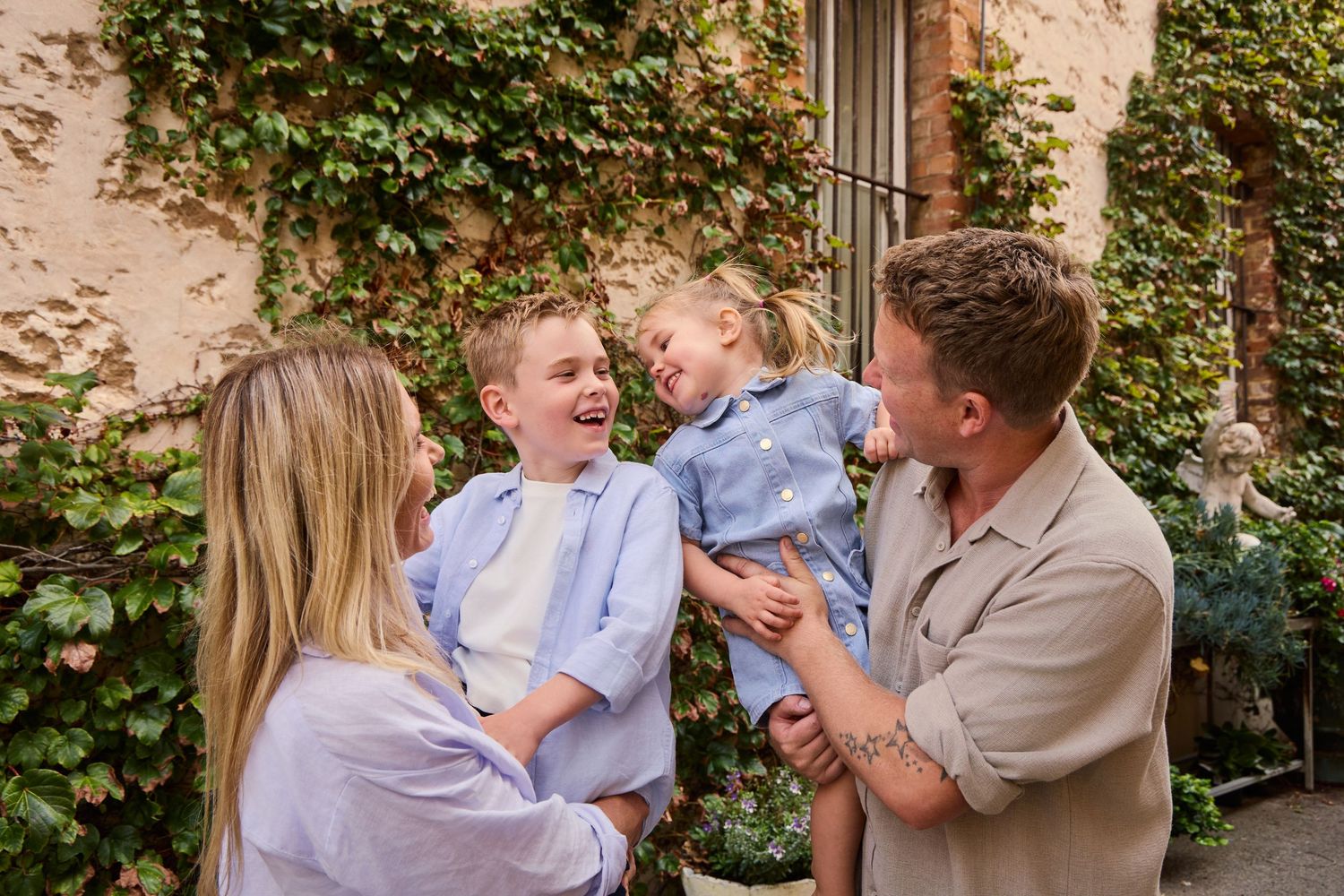 Happy family with two young boys laughing outdoors near ivy-covered stone wall in a European courtyard.