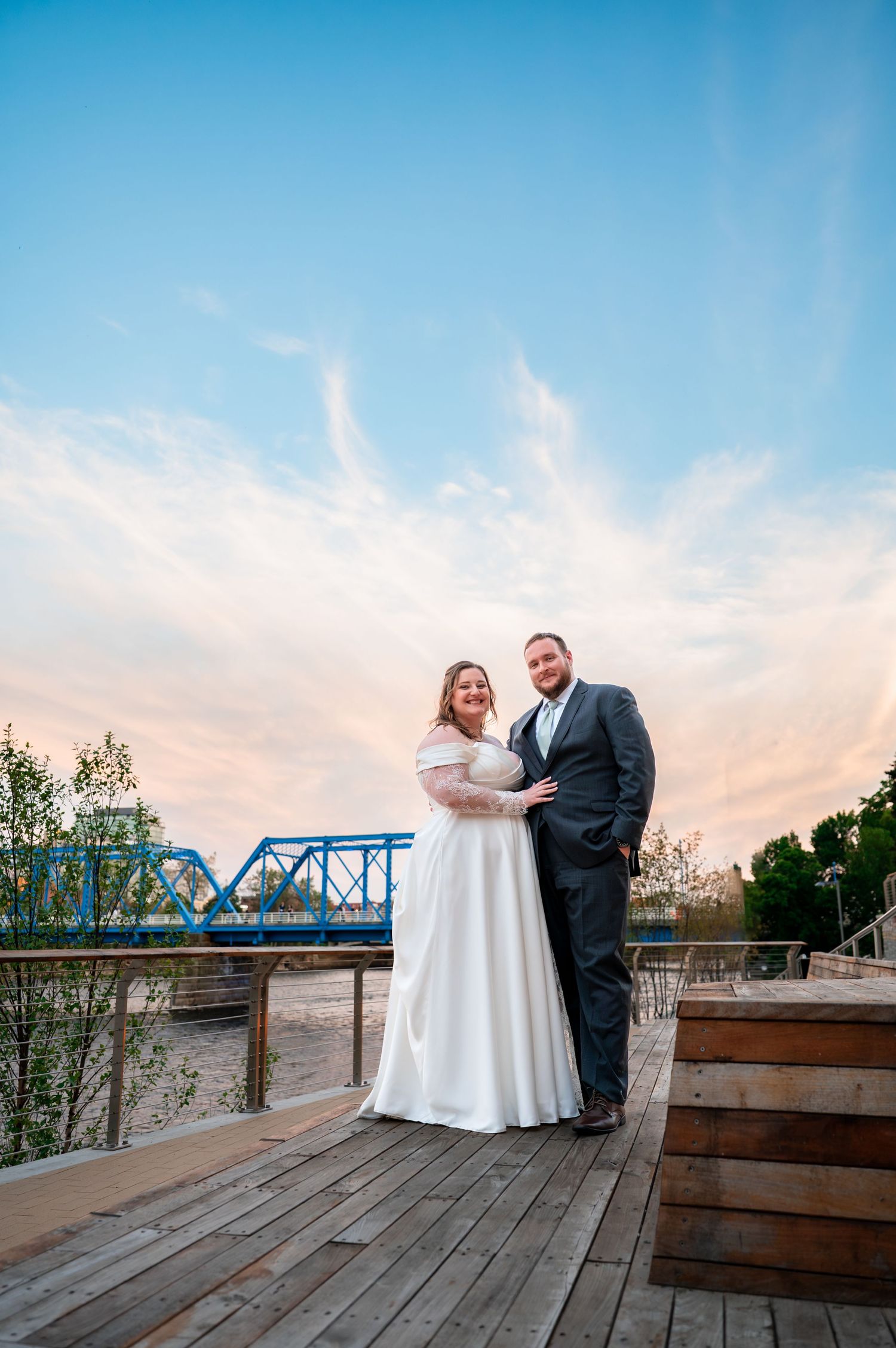 Bride and groom posing on wooden deck at sunset with blue bridge backdrop on their wedding day.