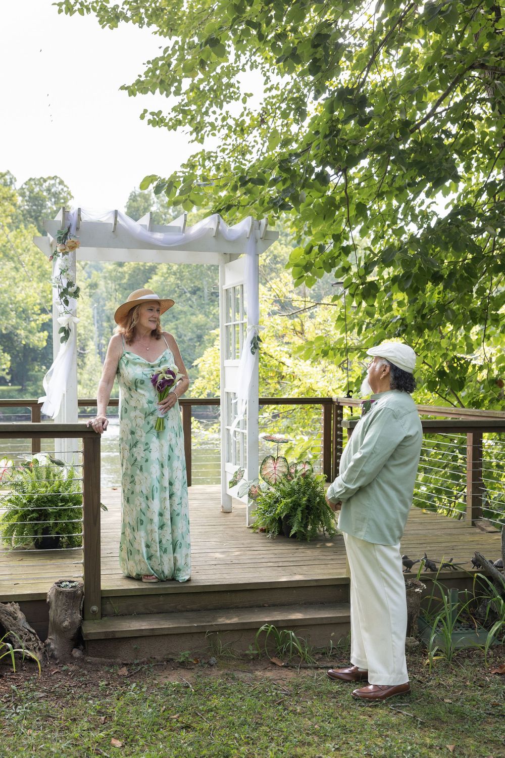 Elderly couple in elegant attire exchange vows on a decorated outdoor deck surrounded by lush green trees.