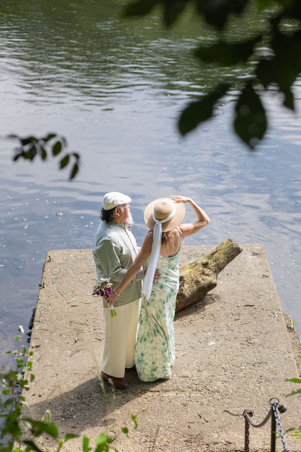 Couple standing on a stone dock by a serene lake, woman in floral dress holding hat, man holding flowers.