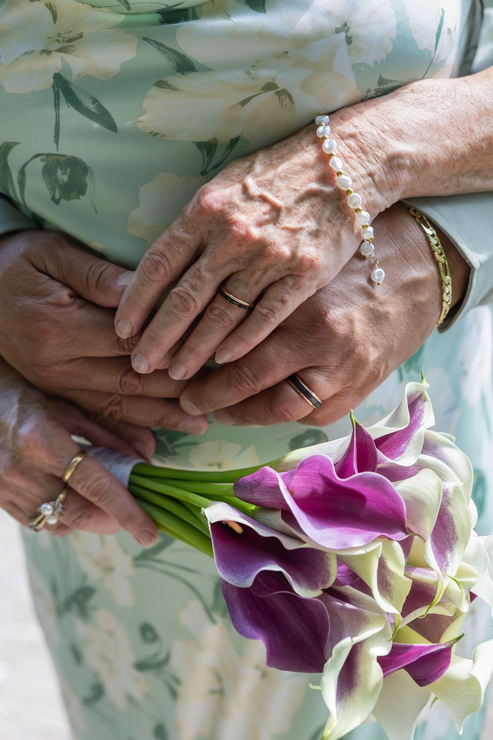 Elderly couple holding hands with wedding rings, pearl bracelet, and purple calla lily bouquet in a floral dress.