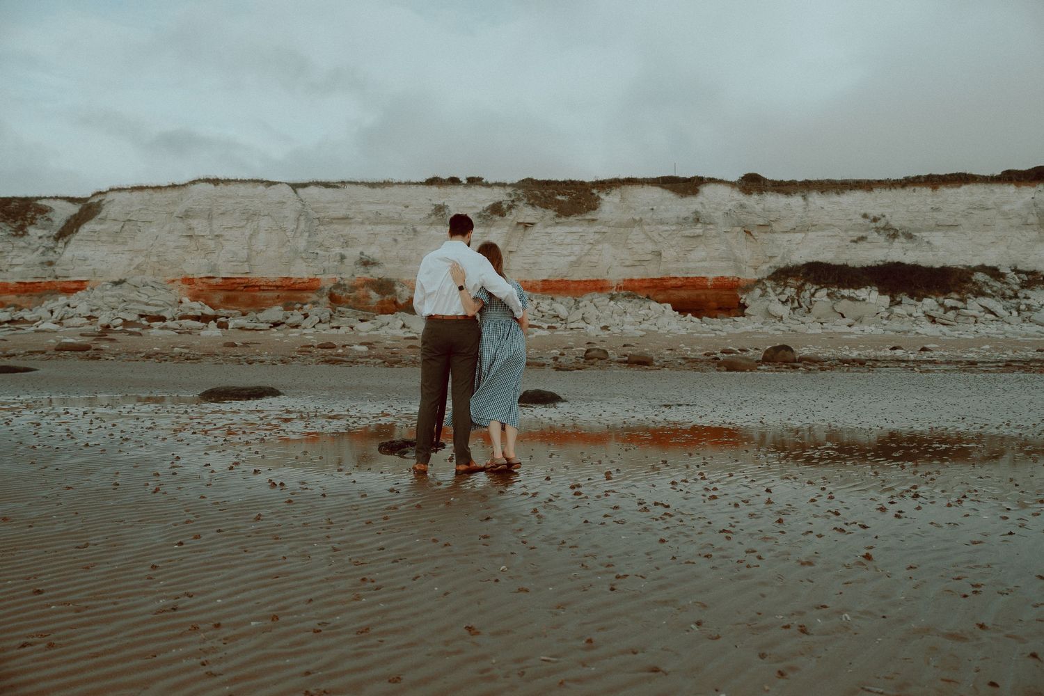 Couple embracing on a wet beach with dramatic white and red striped cliffs in the background on an overcast day.