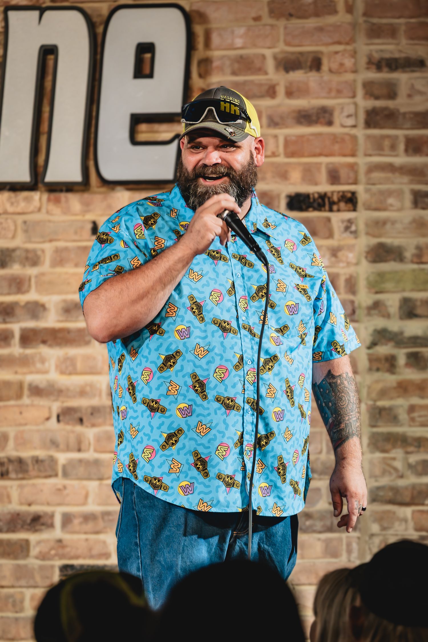 Graeson Mcgaha Bearded male comedian in blue tropical shirt performs stand-up comedy on brick-walled stage holding microphone.