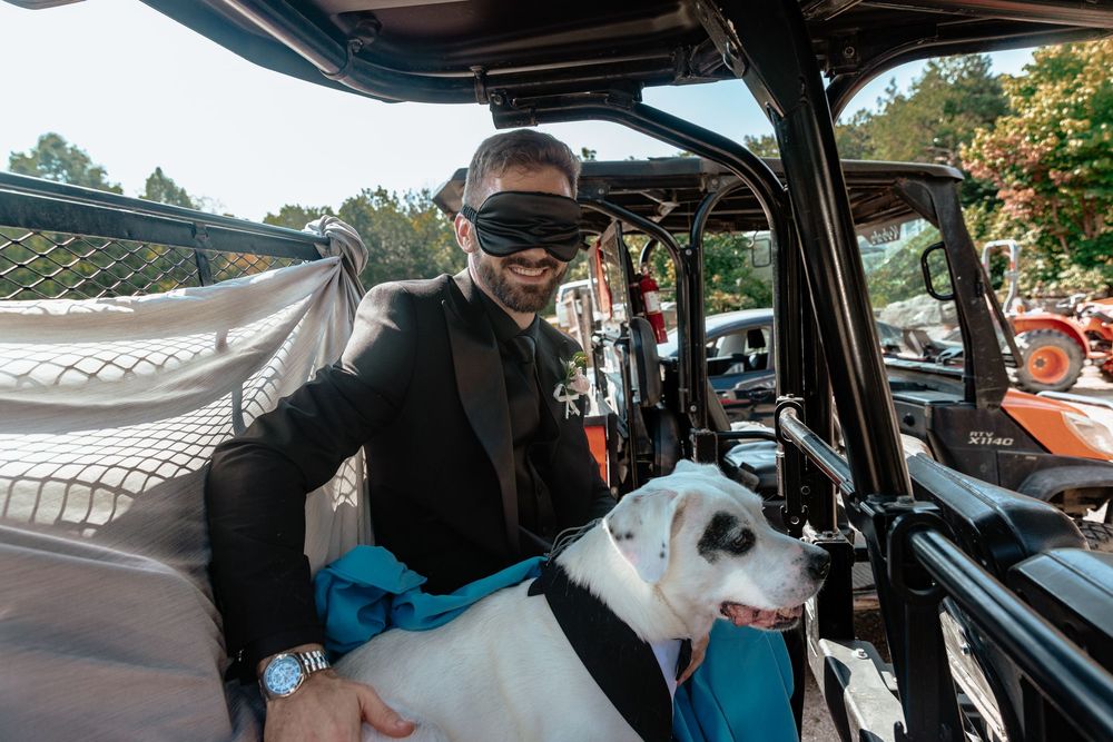 Man in black tuxedo and blindfold smiling while holding a white dog in a decorated vehicle.