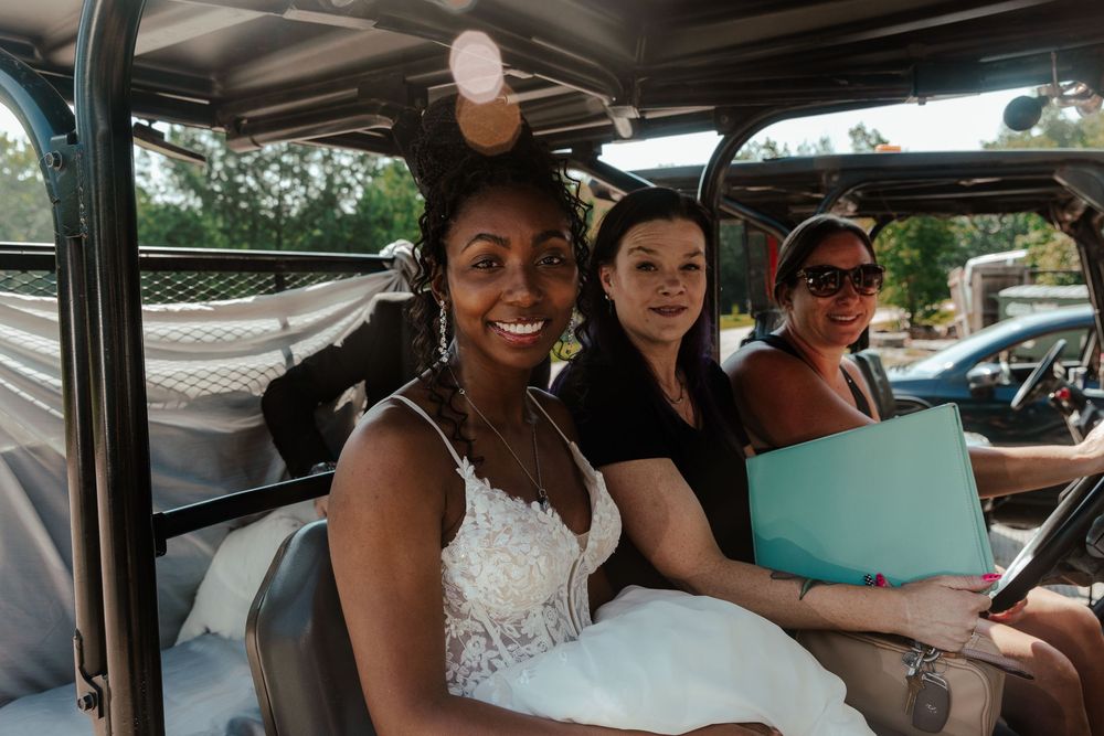 Bride in white lace dress smiles in vintage vehicle with two guests, outdoor wedding celebration.
