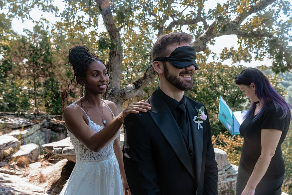 Bride in white lace gown adjusts groom's black blindfold outdoors, as a woman in black watches nearby.