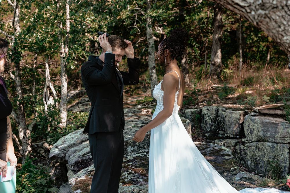 Bride and groom share a romantic first look moment outdoors among trees and rocks at a woodland wedding ceremony.