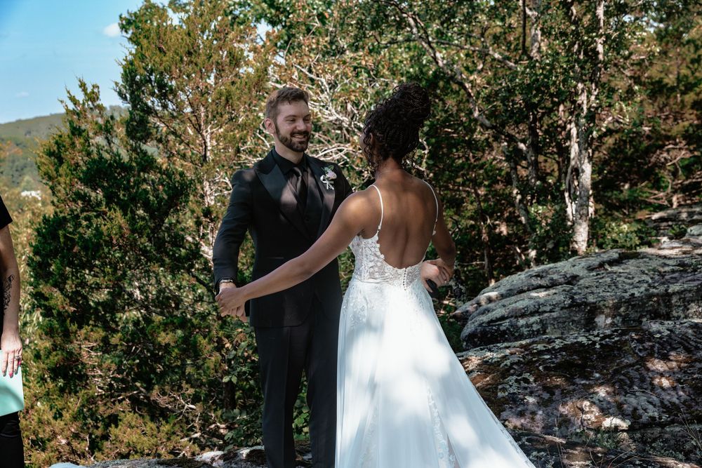 Couple holding hands during outdoor wedding ceremony on rocky terrain surrounded by trees.