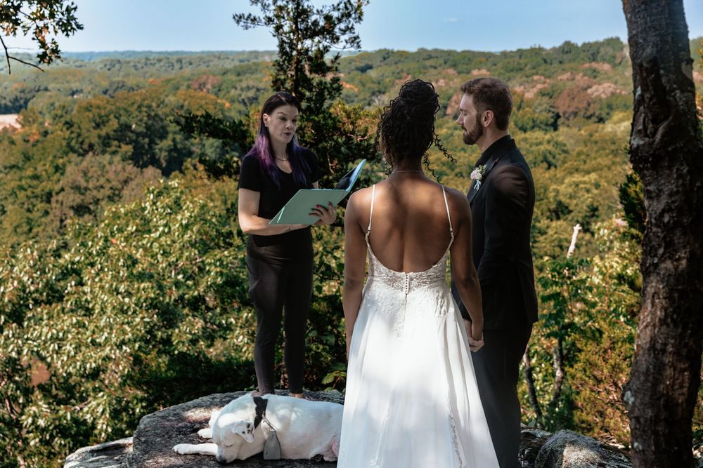Outdoor wedding ceremony on rocky overlook with forest views, officiant reading from book while couple stands facing her.