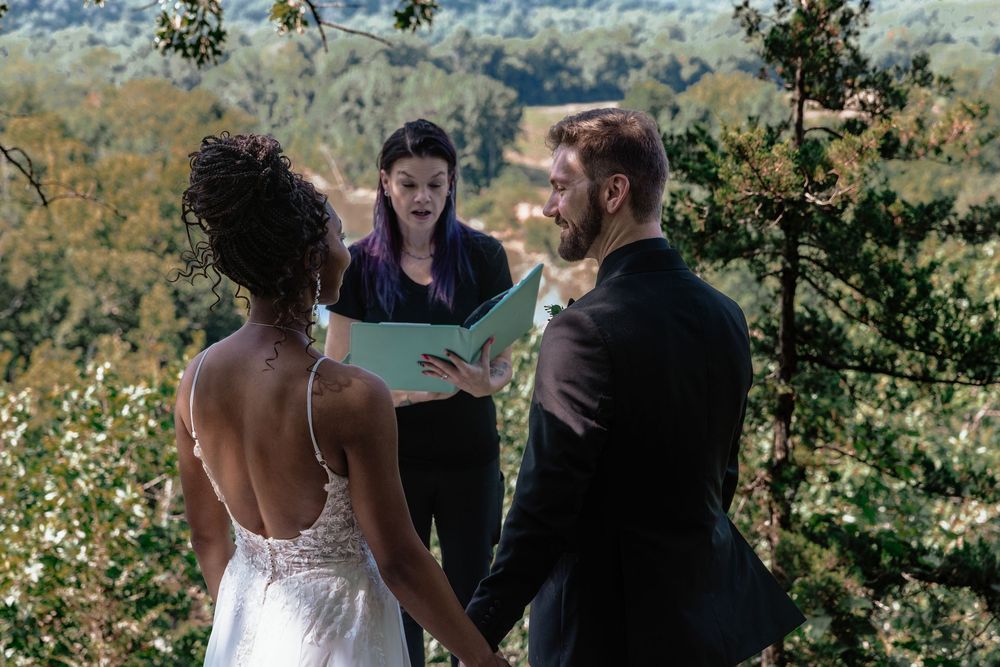 Outdoor wedding ceremony with couple holding hands before officiant, lush green forest backdrop.