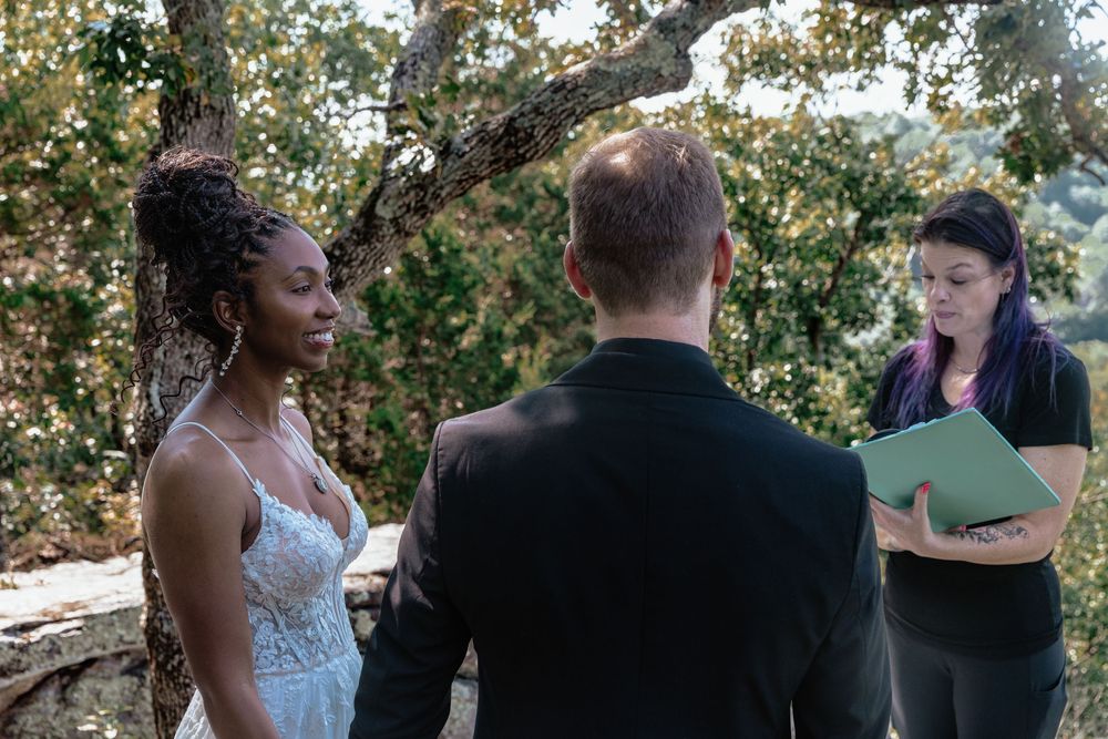 Interracial couple exchanging wedding vows outdoors with officiant holding green folder amid lush tree backdrop.