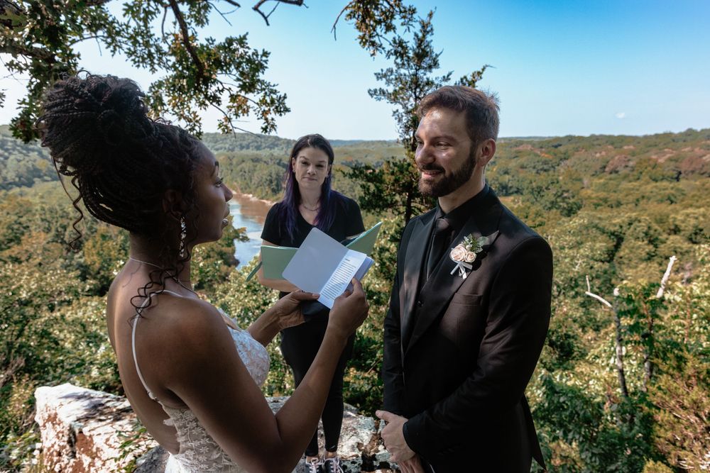 Bride reads vows to groom during outdoor wedding ceremony with scenic forest and river views.