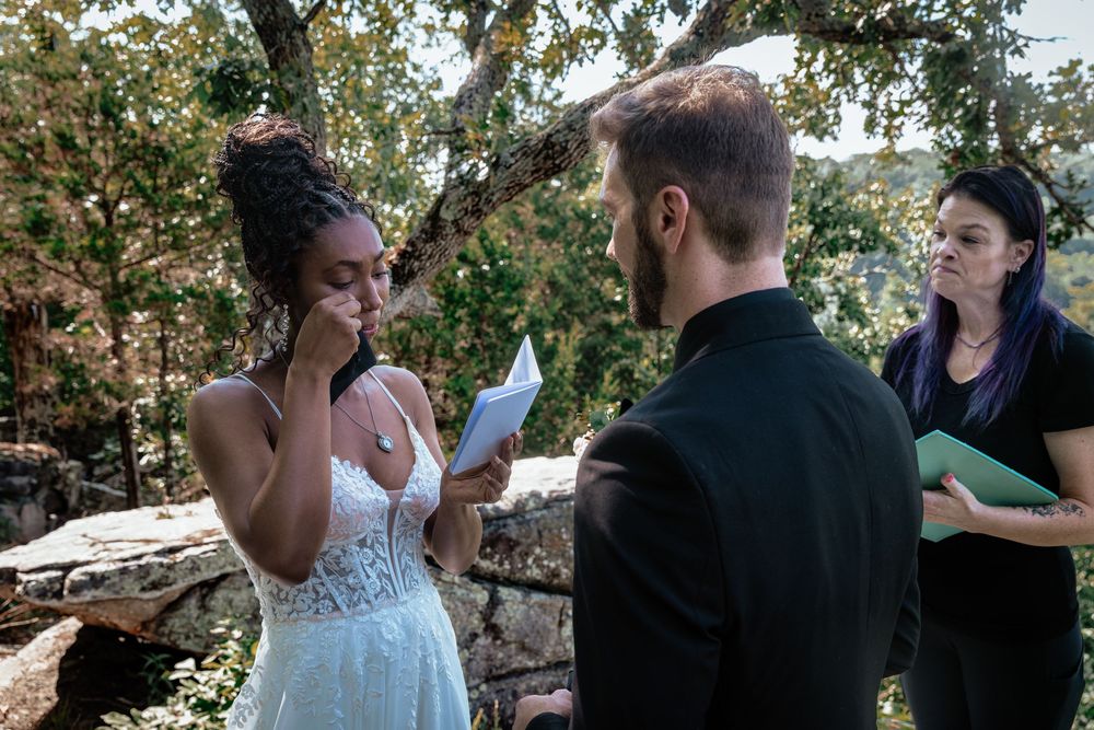 Emotional bride wipes tears reading vows to groom at outdoor woodland wedding ceremony.
